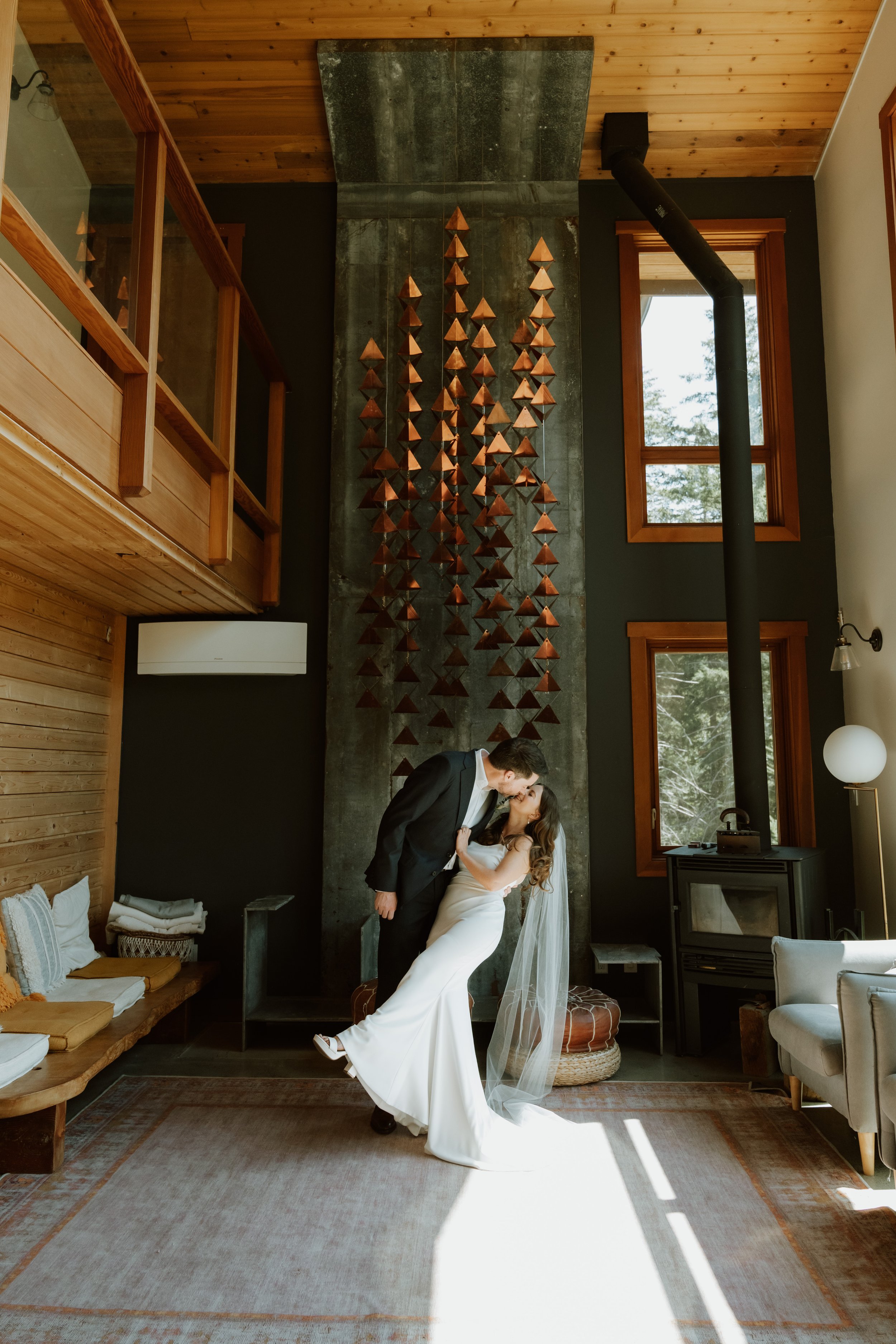 A bride and groom sharing a romantic moment inside a rustic, cozy room with wooden walls, a stone fireplace, and large windows showing trees outside. The bride is wearing a white wedding dress and veil, and the groom is dressed in a dark suit, leanin