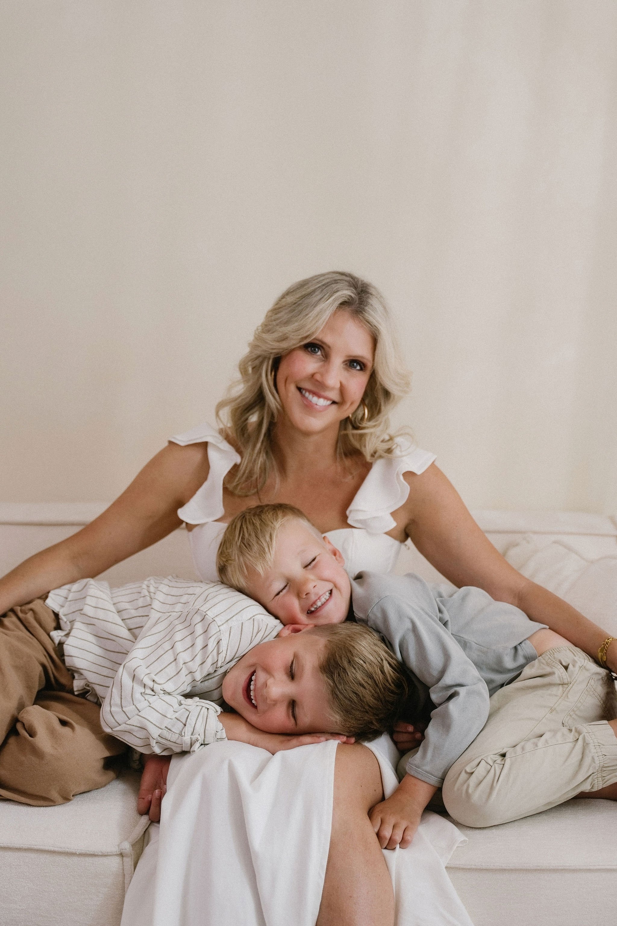 A woman with blonde hair smiling with three young boys, lying on a bed, relaxing and enjoying each other's company.
