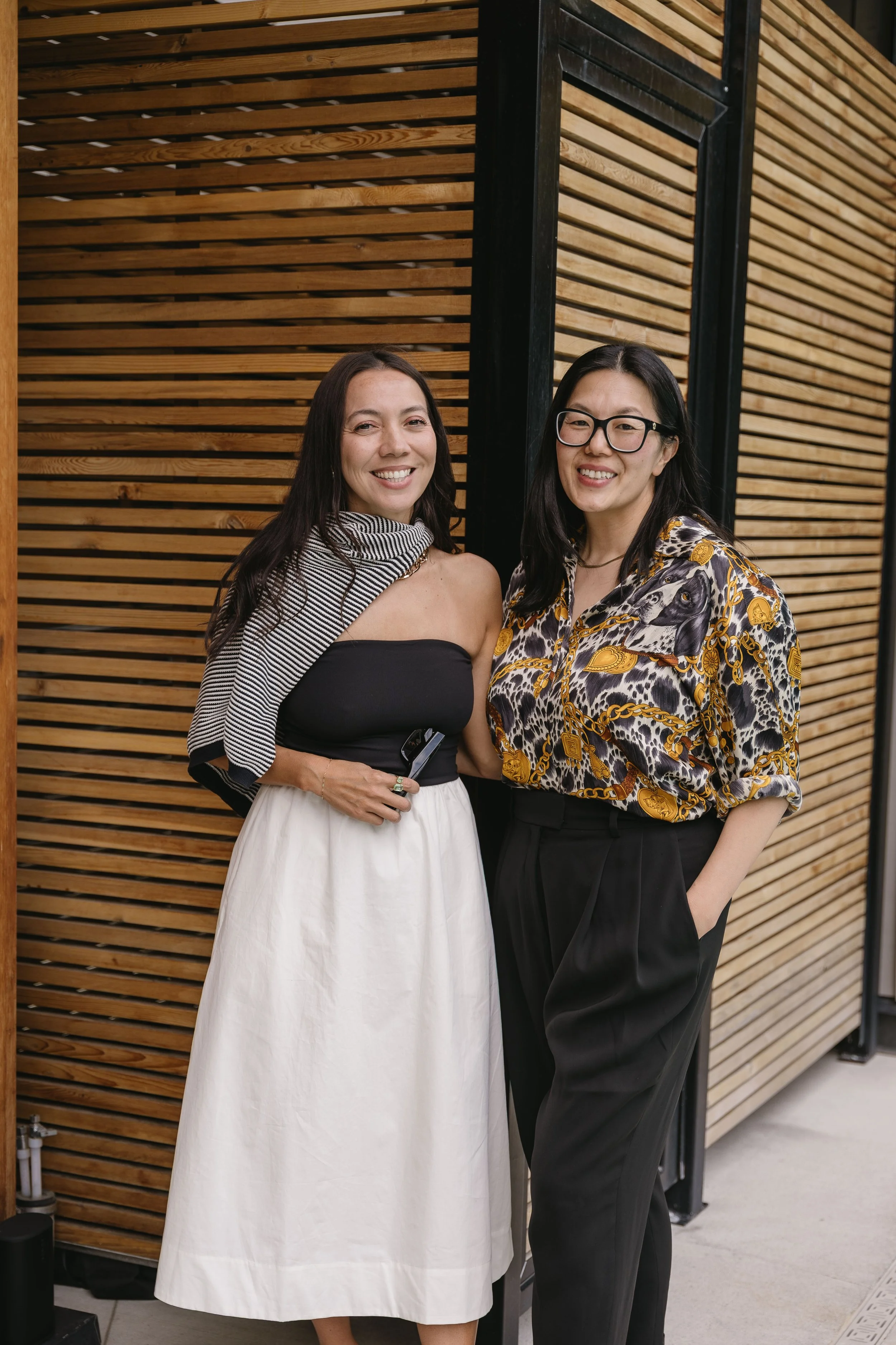 Two women standing side by side in front of a wooden panel wall, smiling at the camera. One woman is wearing a strapless black top with a white skirt and a striped shawl, and the other is dressed in a patterned shirt with animal print and black high-