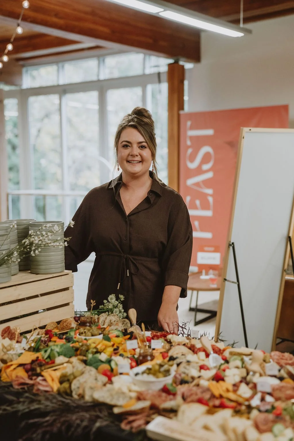 A woman standing behind a table with a variety of cheeses, vegetables, and crackers at a catering or event display inside a room with large windows and wooden ceiling beams.