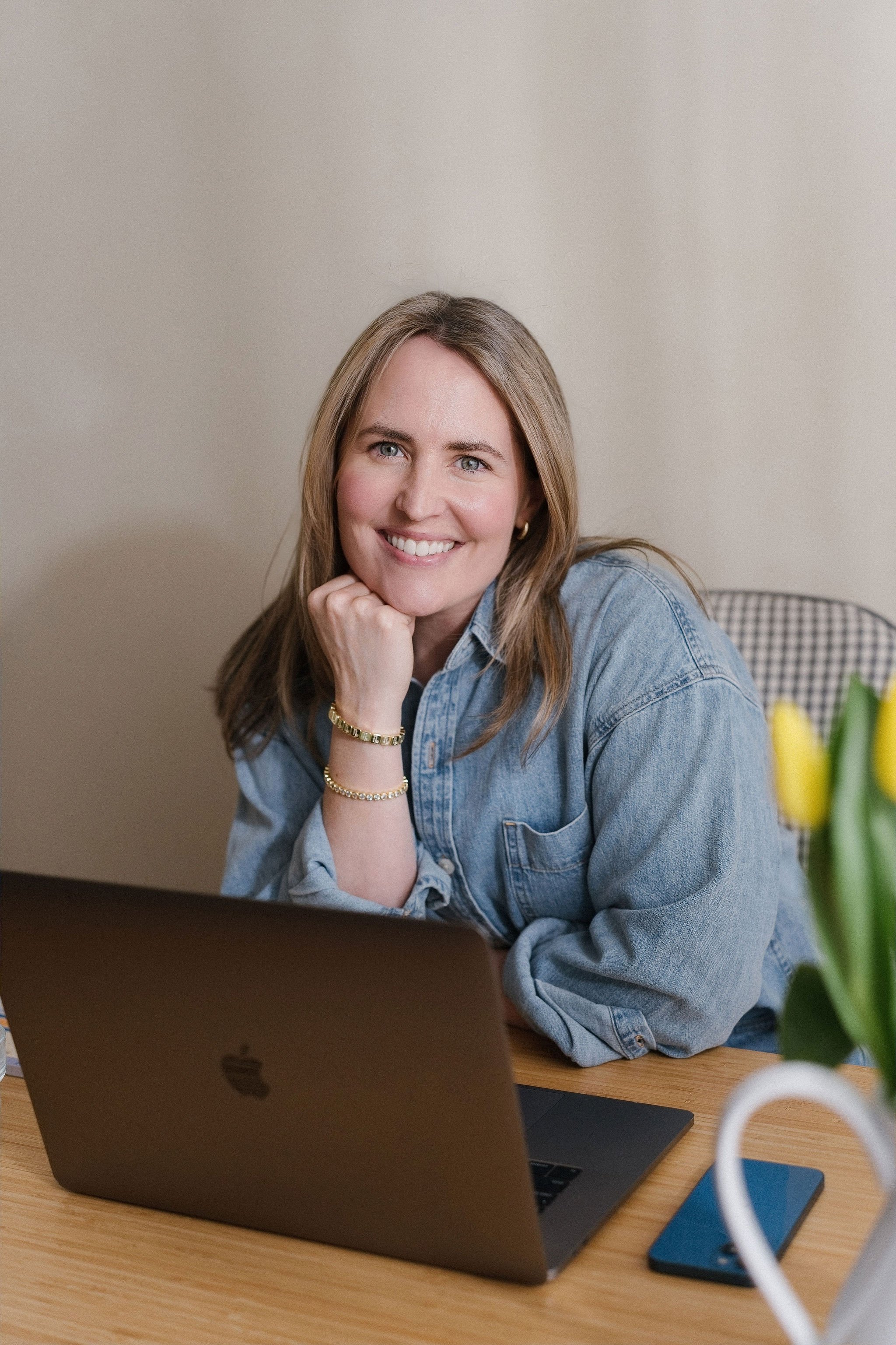 A smiling woman with blonde hair, wearing a denim shirt and gold jewelry, sitting at a wooden desk with a silver MacBook laptop and a potted plant in the foreground.