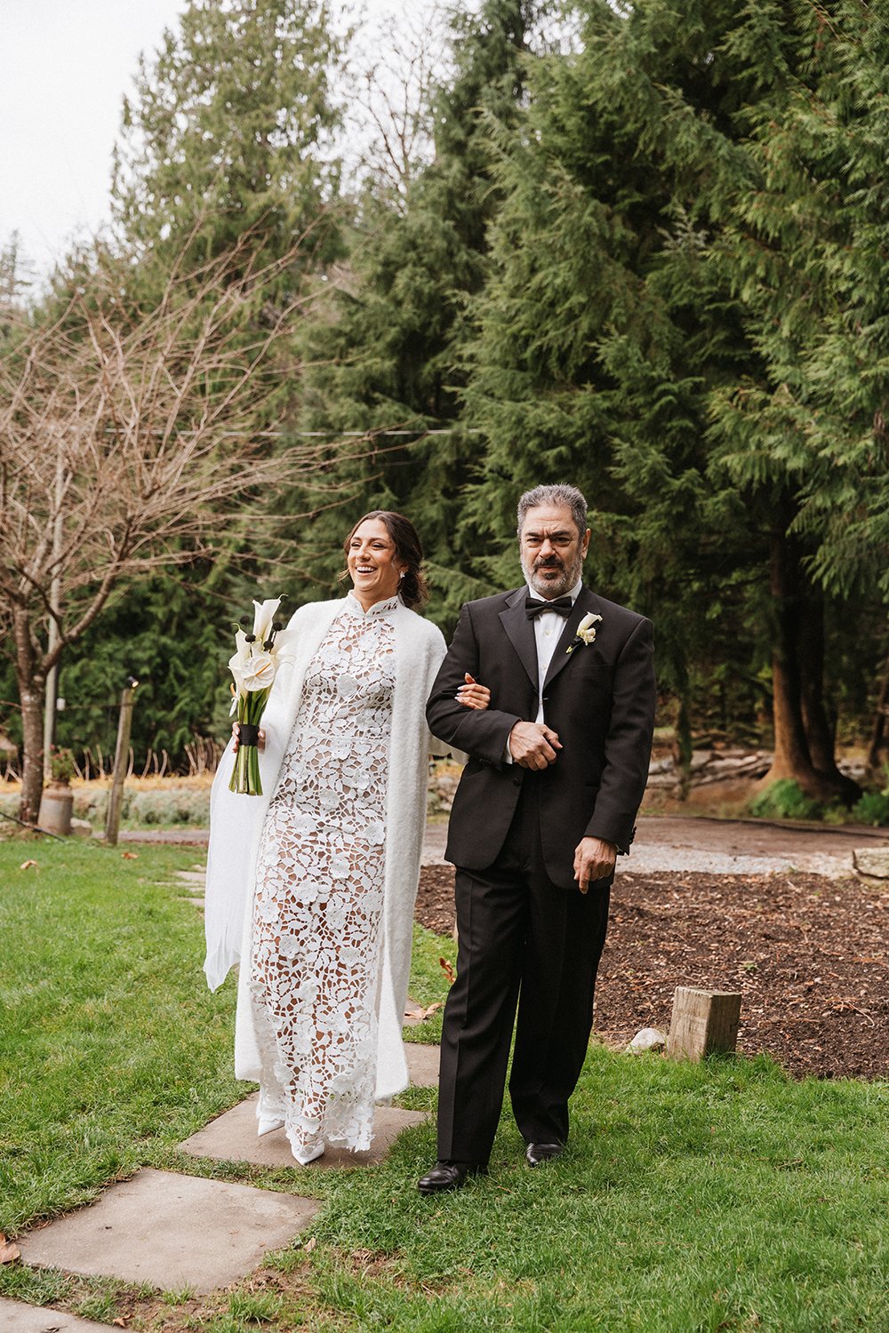 A bride and a man, possibly her father, walking outdoors on a wedding day. The bride is dressed in a white lace gown and holding a bouquet of white flowers, and the man is wearing a black tuxedo.