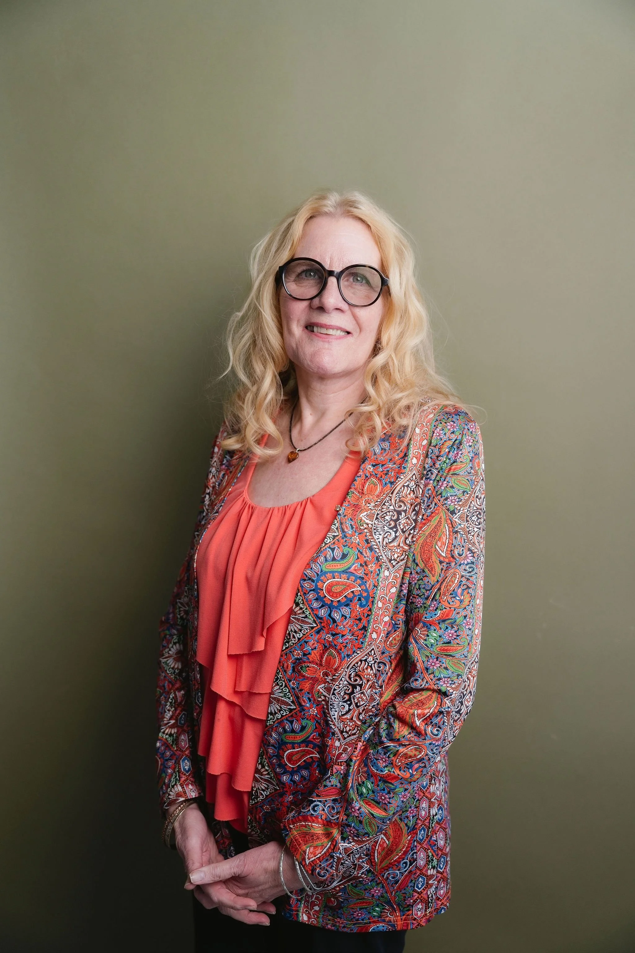 A woman with long, wavy blonde hair and glasses, wearing a colorful paisley-patterned jacket over an orange top with a ruffled front, standing against a plain, solid green background.