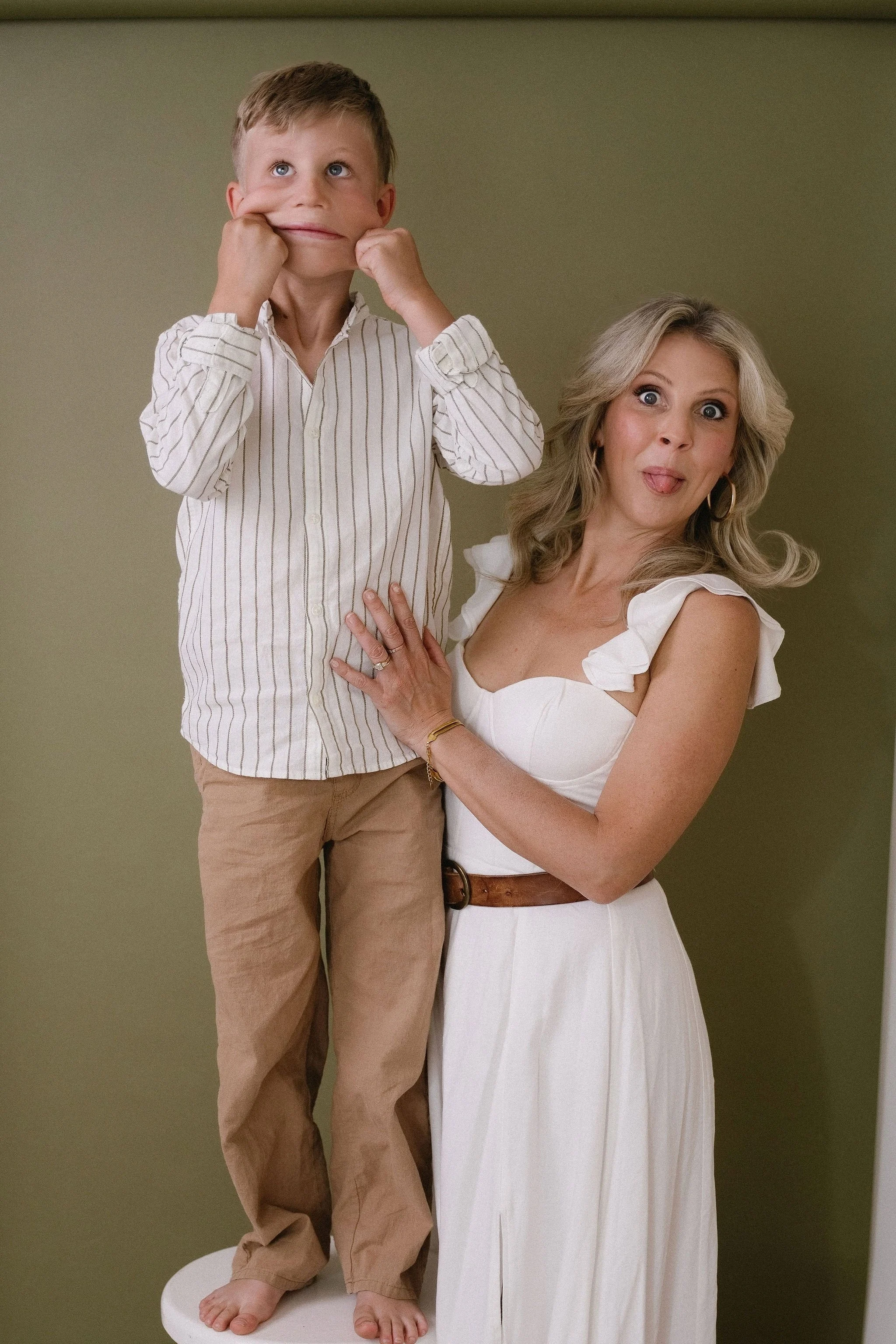 A woman with gray hair and wearing a white dress with ruffled sleeves is playfully sticking her tongue out and holding a young boy who is standing on a white pedestal. The boy is making a funny face by pulling his cheeks and looking up with his mouth