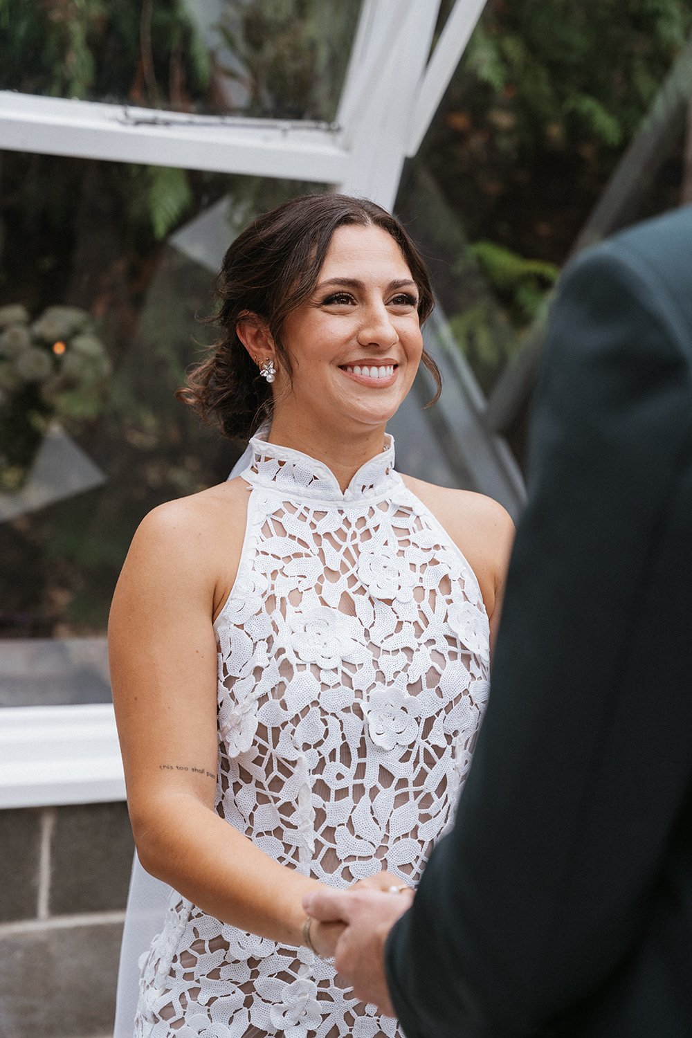 A smiling woman in a white lace dress is holding hands with a man during a wedding ceremony.