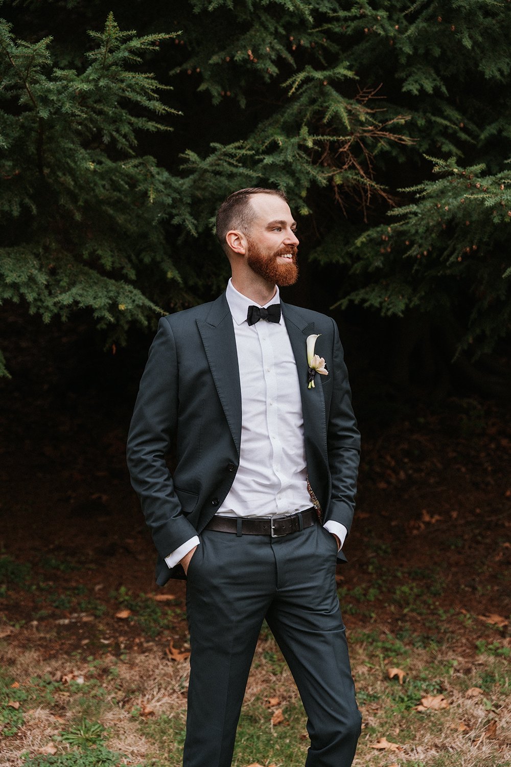 A man dressed in a tuxedo with a bow tie and boutonniere, standing outdoors in front of dense green trees, smiling and looking off to his right.