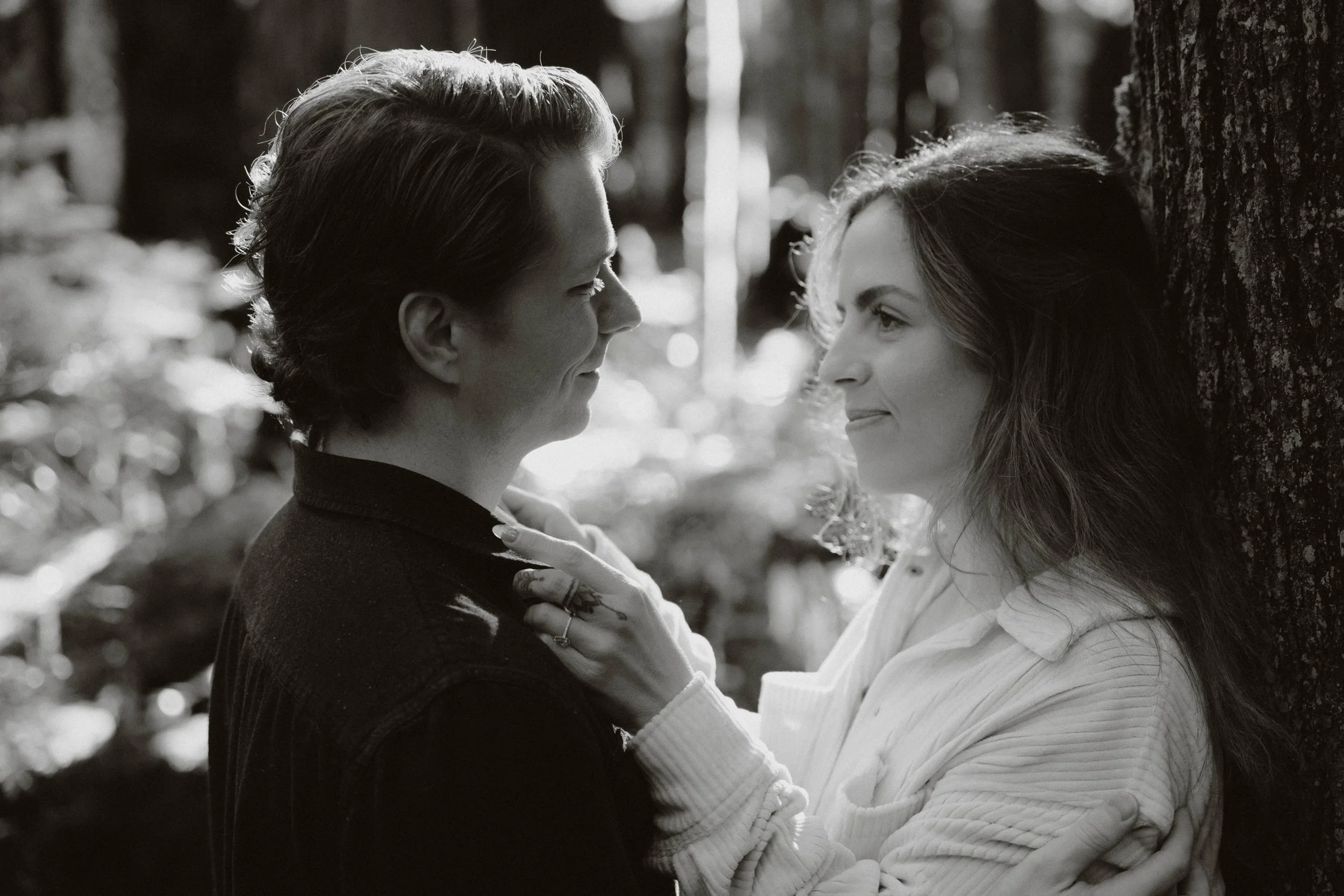 A black and white photo of a man and a woman facing each other closely outdoors among trees. The man is wearing a dark suit, and the woman has long hair and is wearing a white sweater. They are gazing into each other's eyes with gentle expressions.