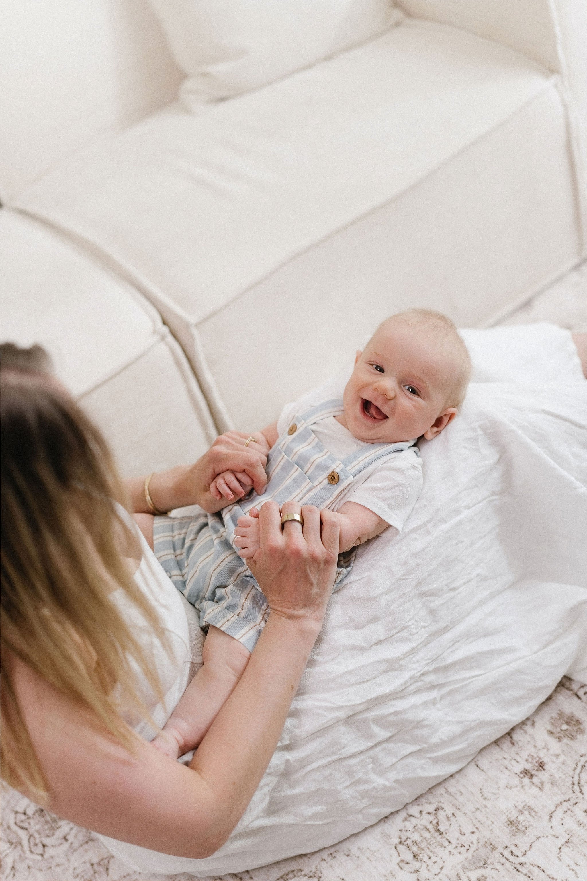A smiling baby lying on a bed, holding hands with a woman, with a white headboard and beige carpet in the background.