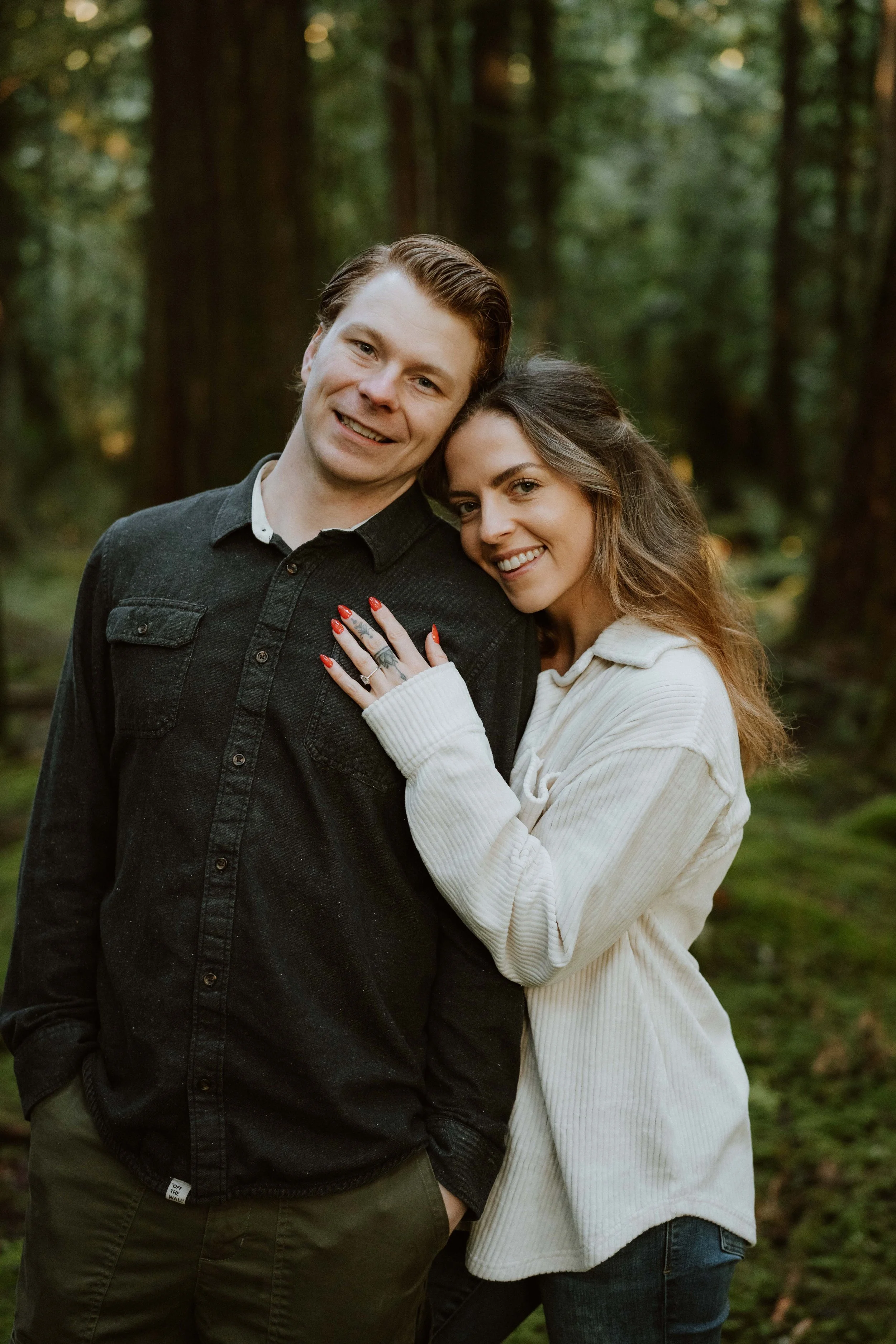 A smiling couple standing close together outdoors in a wooded area, with the woman leaning her head on the man's shoulder.