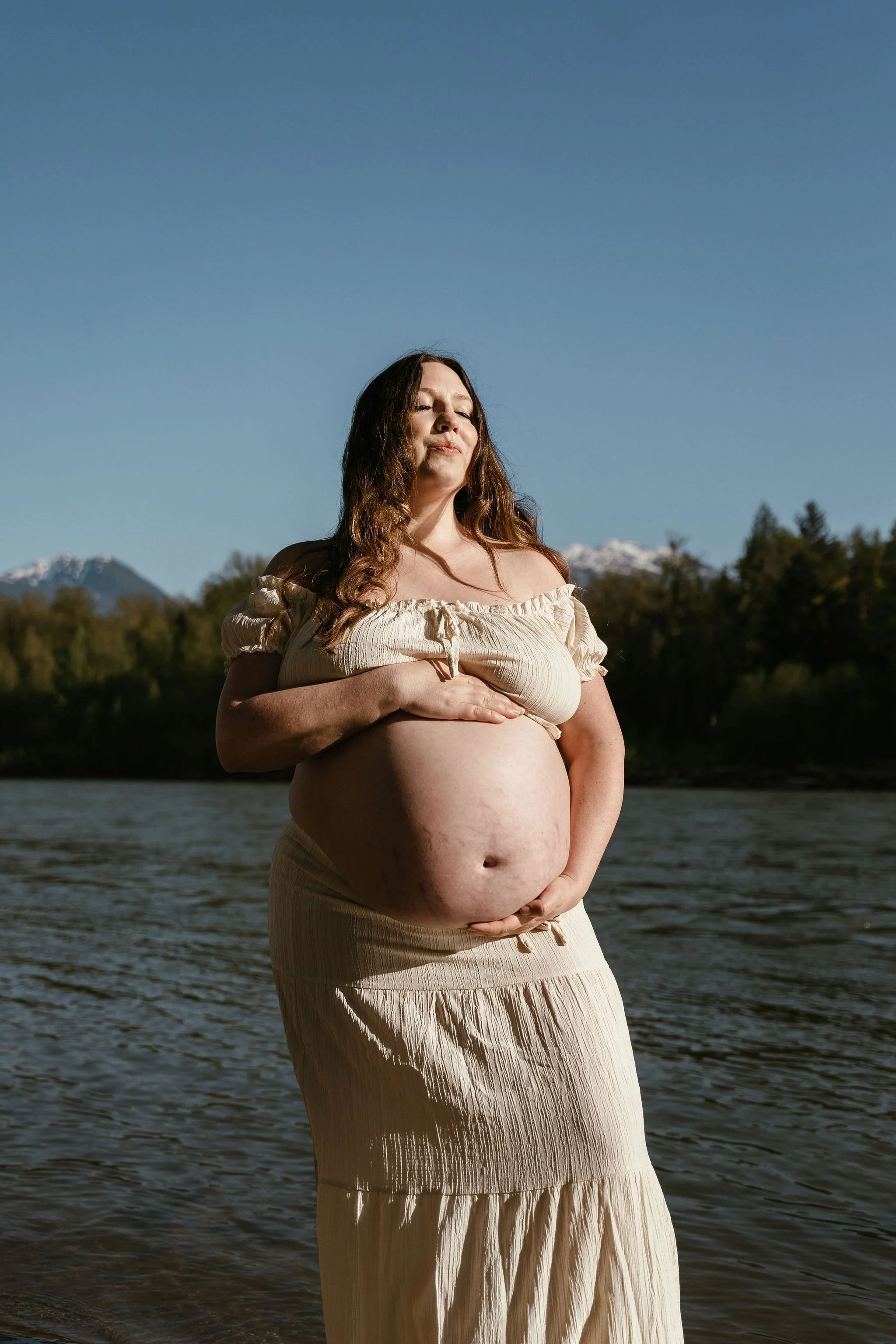 A pregnant woman with long brown hair stands near a river, gently touching her belly, with her eyes closed and a peaceful expression, wearing a beige off-shoulder top and a matching skirt, with snowy mountains and trees in the background under a clea