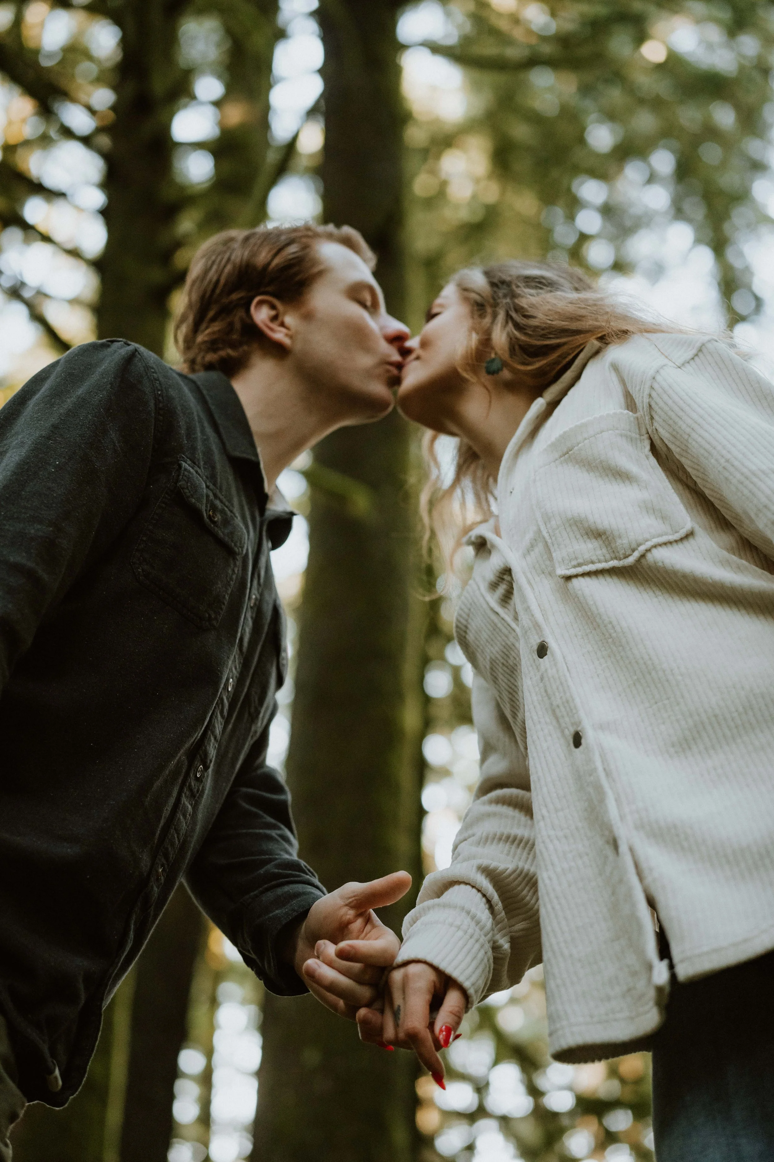 A couple is kissing in a forest, holding hands, with trees and sunlight in the background.