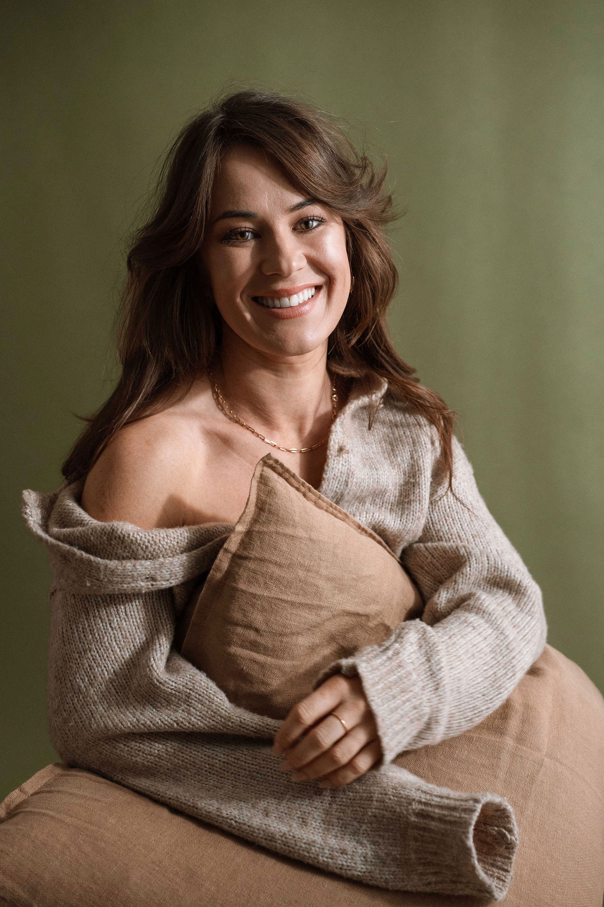 A woman with brown hair wearing a beige sweater, holding a light brown cushion, and sitting in front of a green background, smiling at the camera.