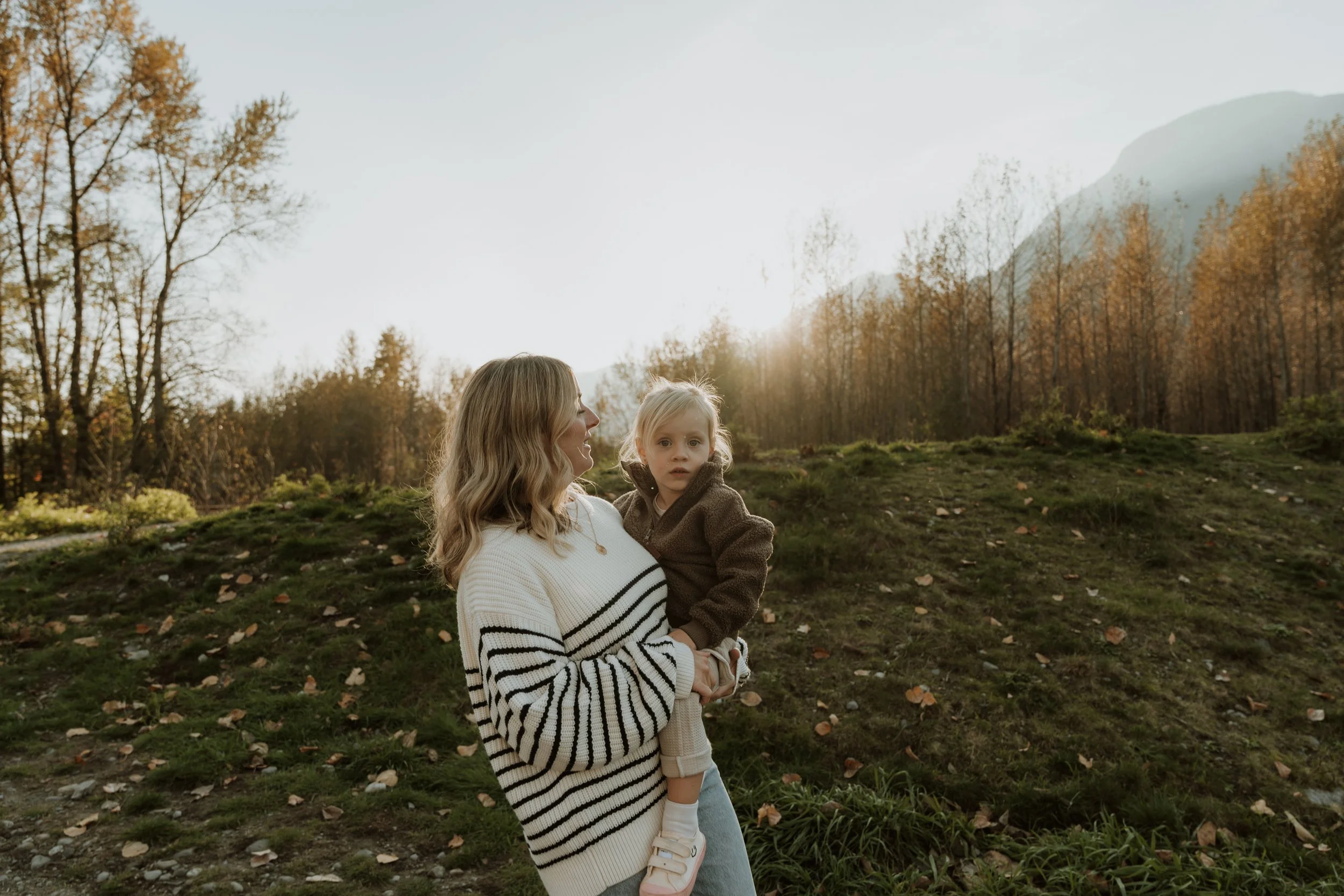 A woman holding a young girl outdoors in a natural setting with trees and mountains in the background during sunset.