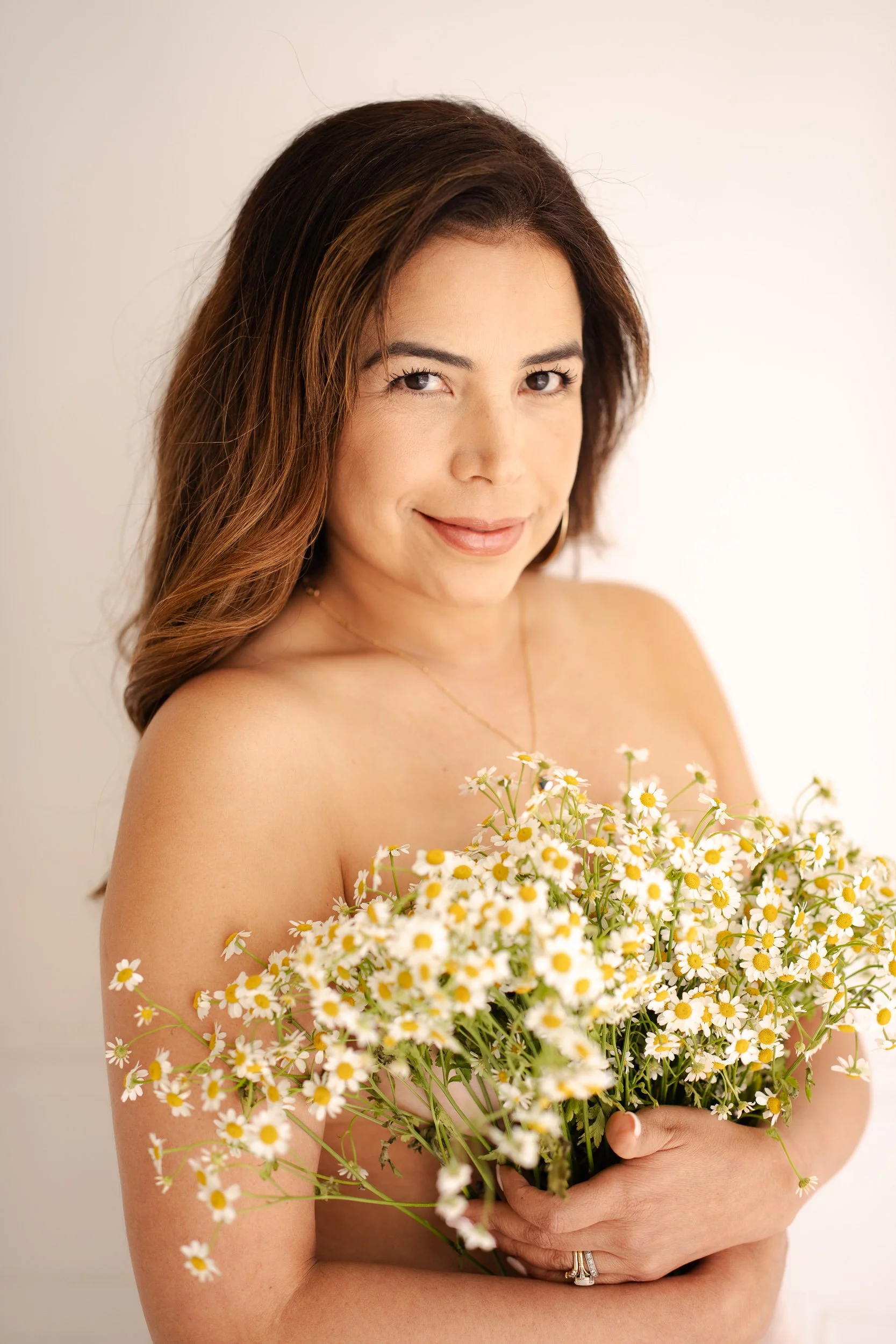 A woman with long, wavy brown hair holds a large bouquet of daisies and smiles softly at the camera.