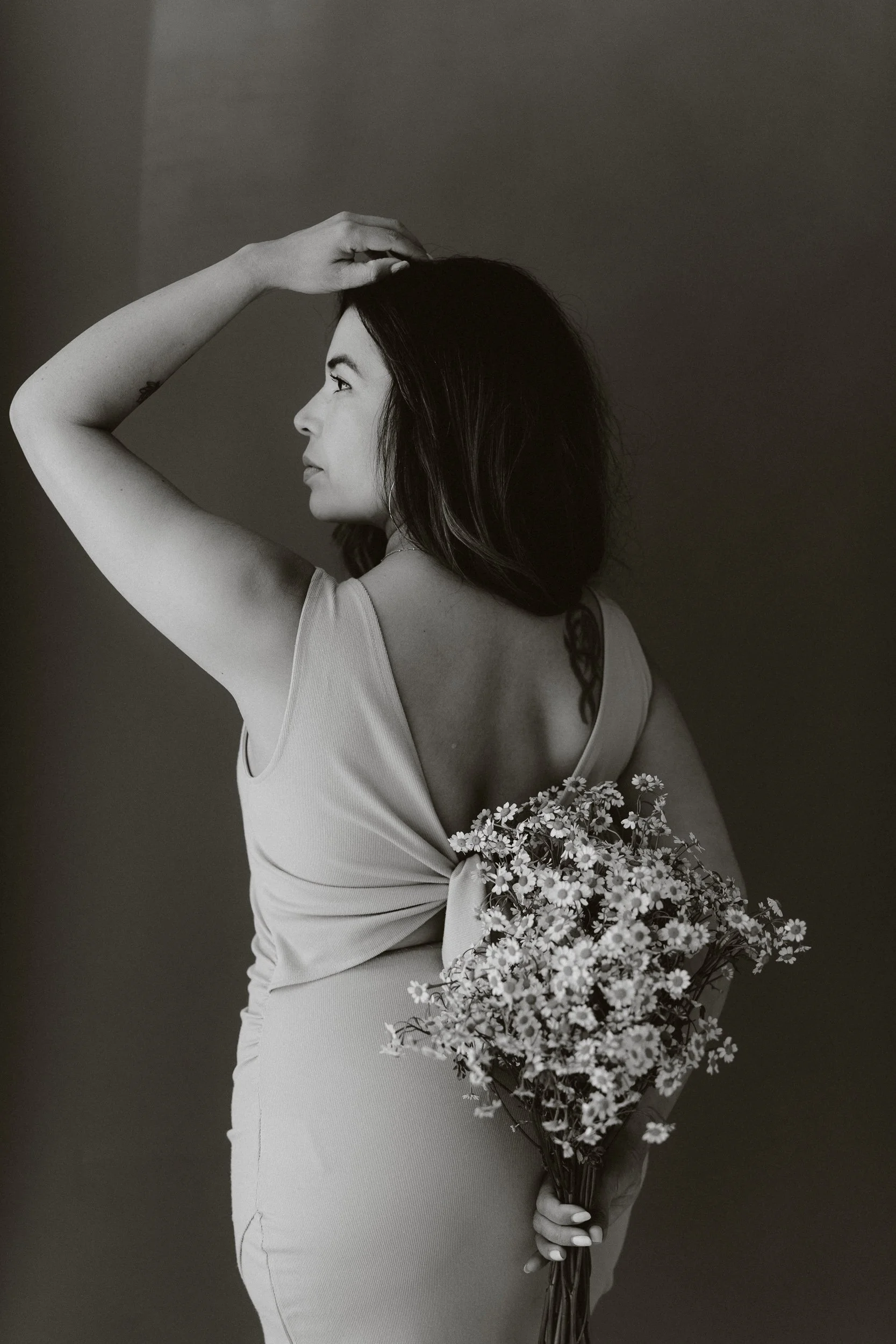 A woman in a sleeveless dress is holding a bouquet of small flowers behind her back, with her left hand and resting her right hand on her head, looking to the left against a plain background.