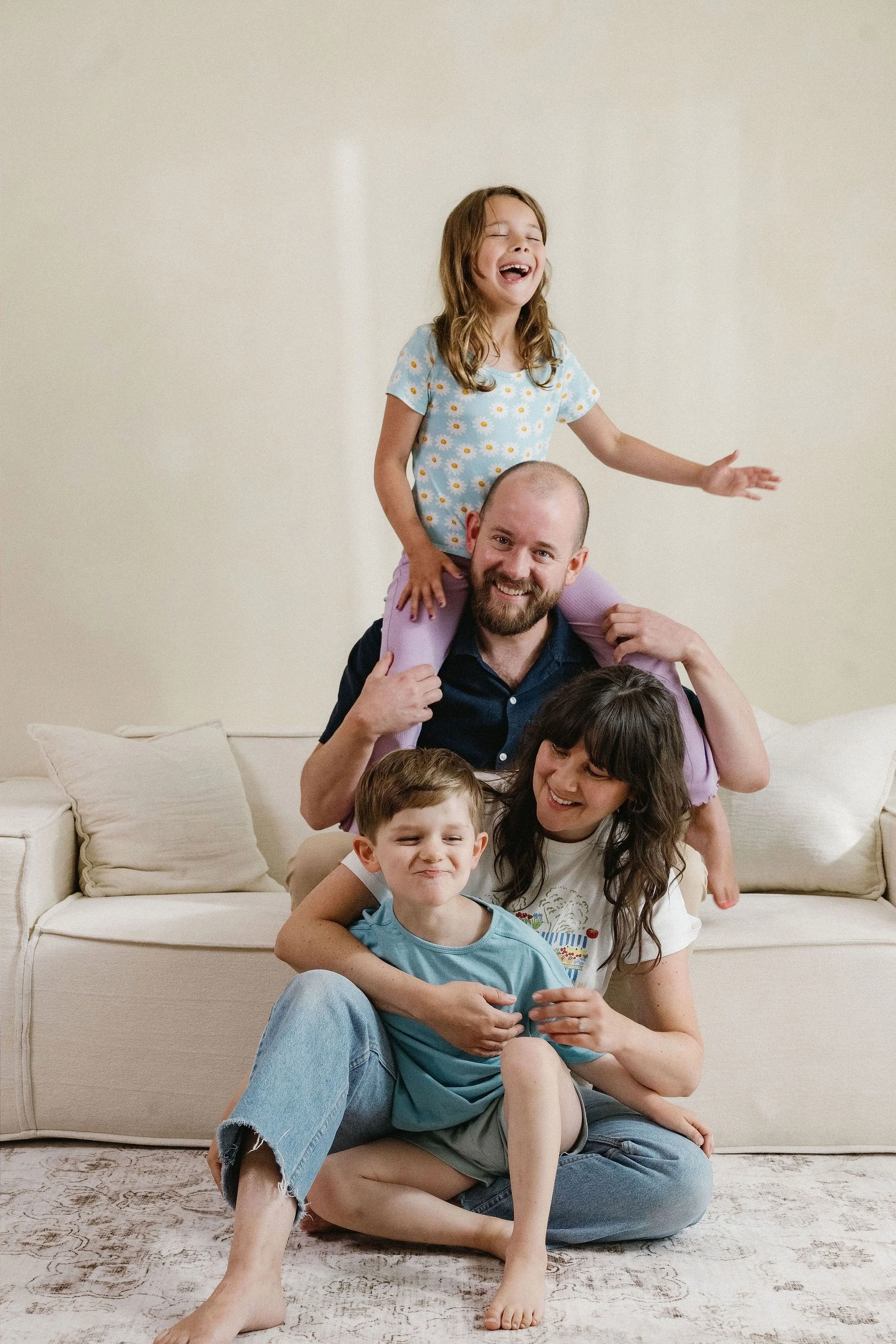 Happy family of six posing on a beige sofa in a living room. A young girl with long hair is sitting on a man's shoulders, laughing. The man with a beard is smiling. A woman with dark hair and bowl cut is sitting on the floor in front. A boy and a gir