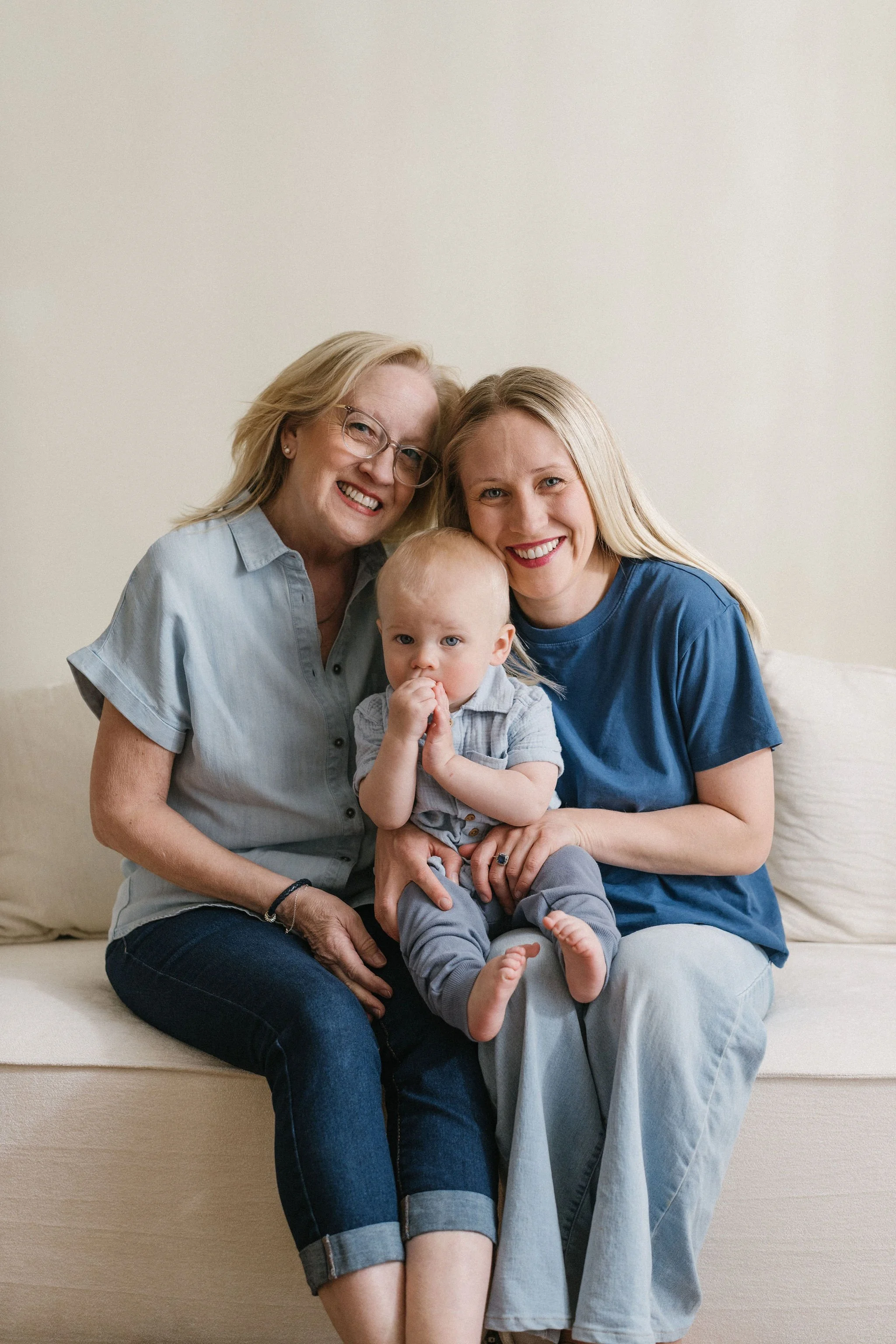 A multigenerational family of three women and a baby sitting together on a beige sofa in a room with a plain light-colored wall, smiling at the camera.