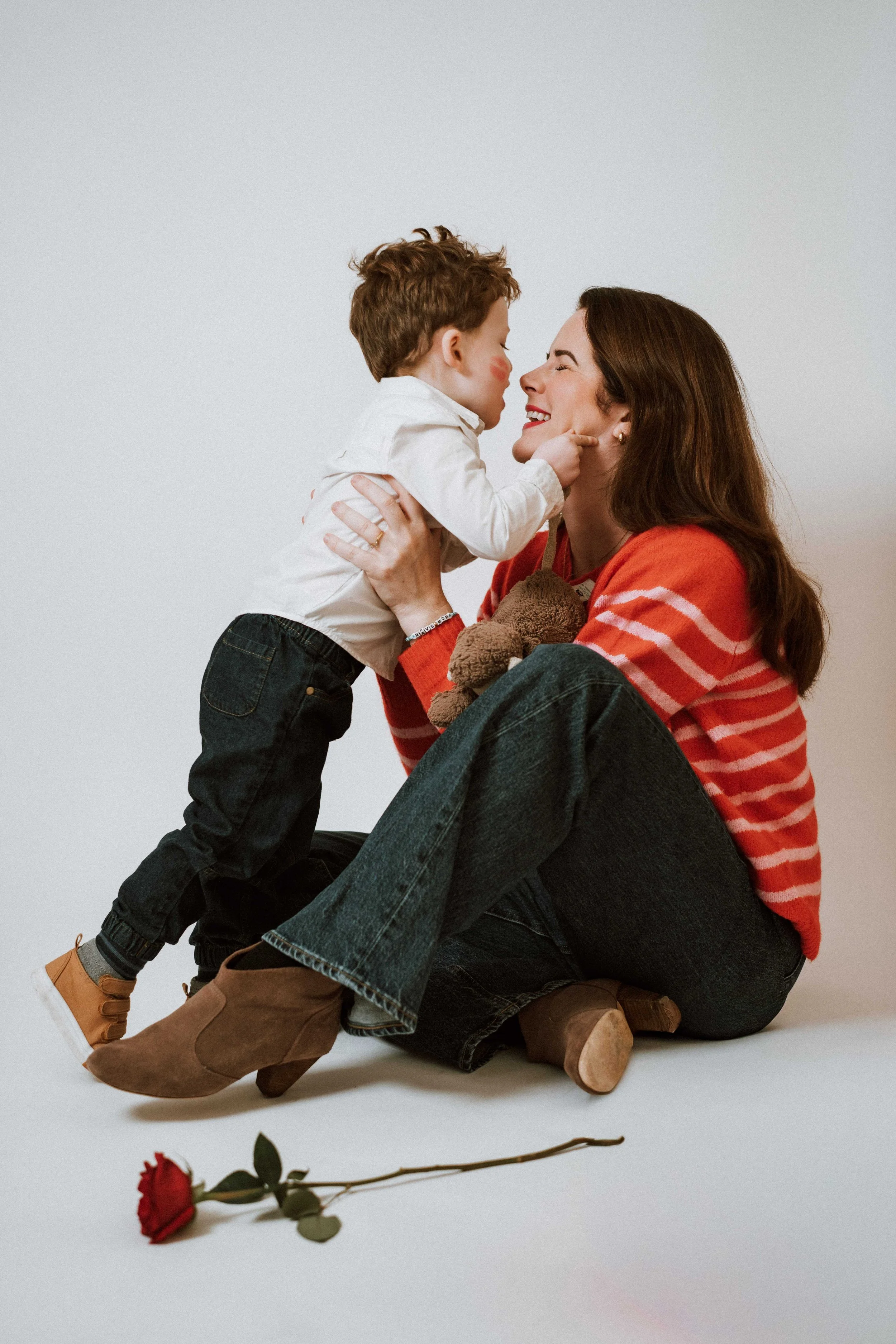 A woman and a young boy sharing a joyful moment, with their faces close together, the woman smiling and the boy with makeup kiss marks on his cheek. The woman is sitting on the floor with her knee bent, wearing a red and pink striped sweater and jean
