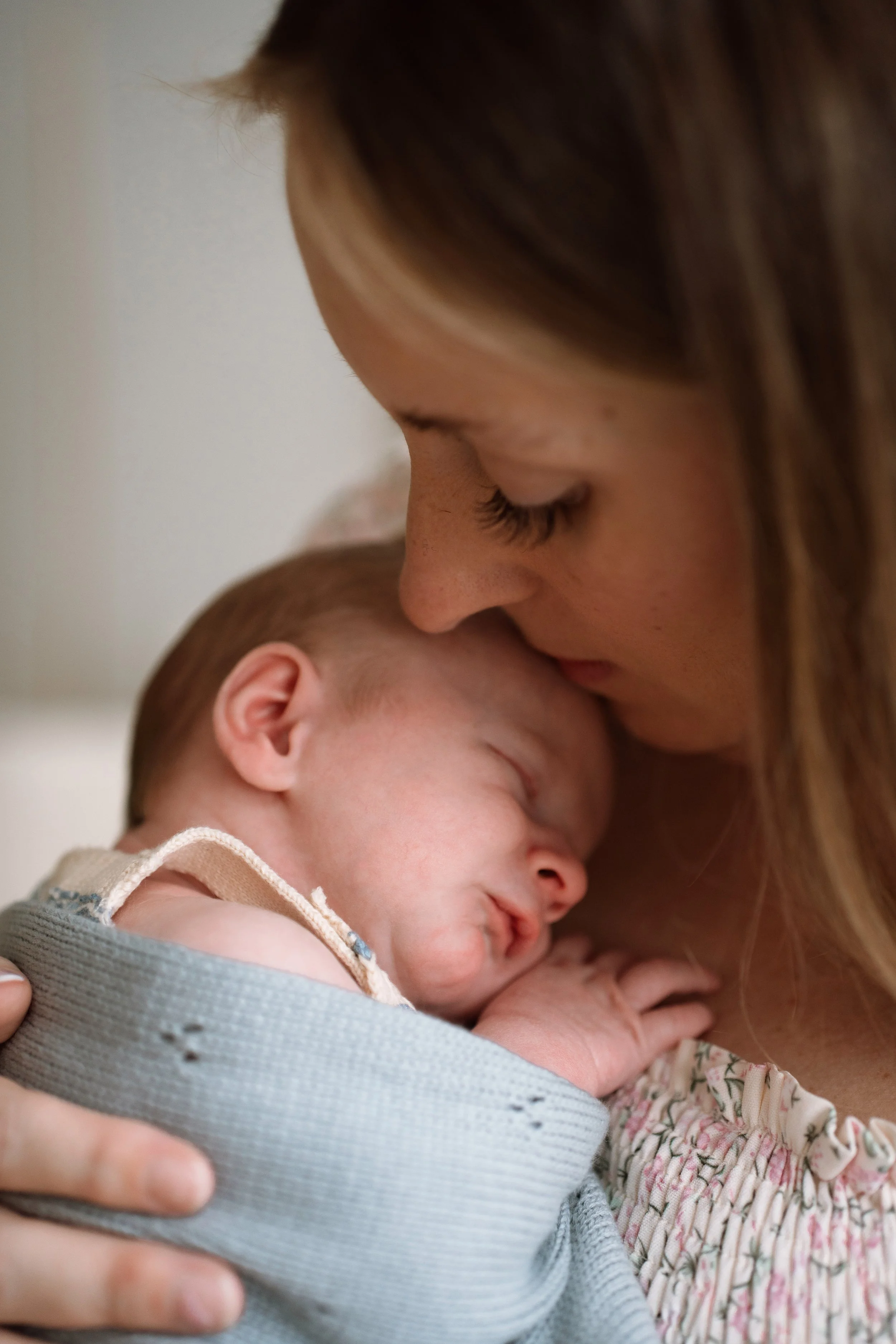 A woman with long, brown hair and closed eyes gently kisses a sleeping baby with light skin, brown hair, and closed eyes, who is swaddled in a blanket.