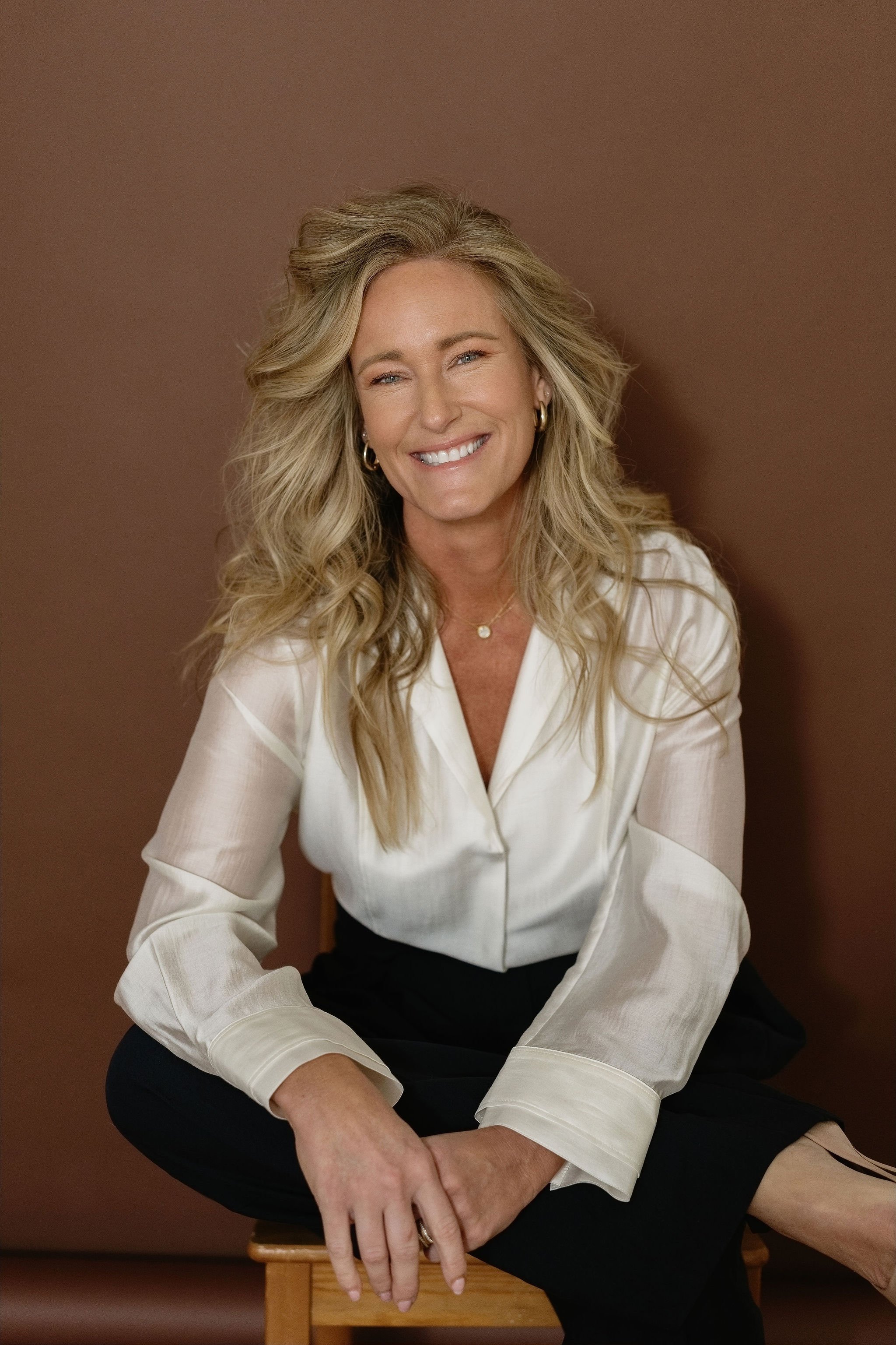 Woman with long wavy blonde hair smiling, wearing a white blouse with rolled-up sleeves, sitting on a wooden stool against a brown background.