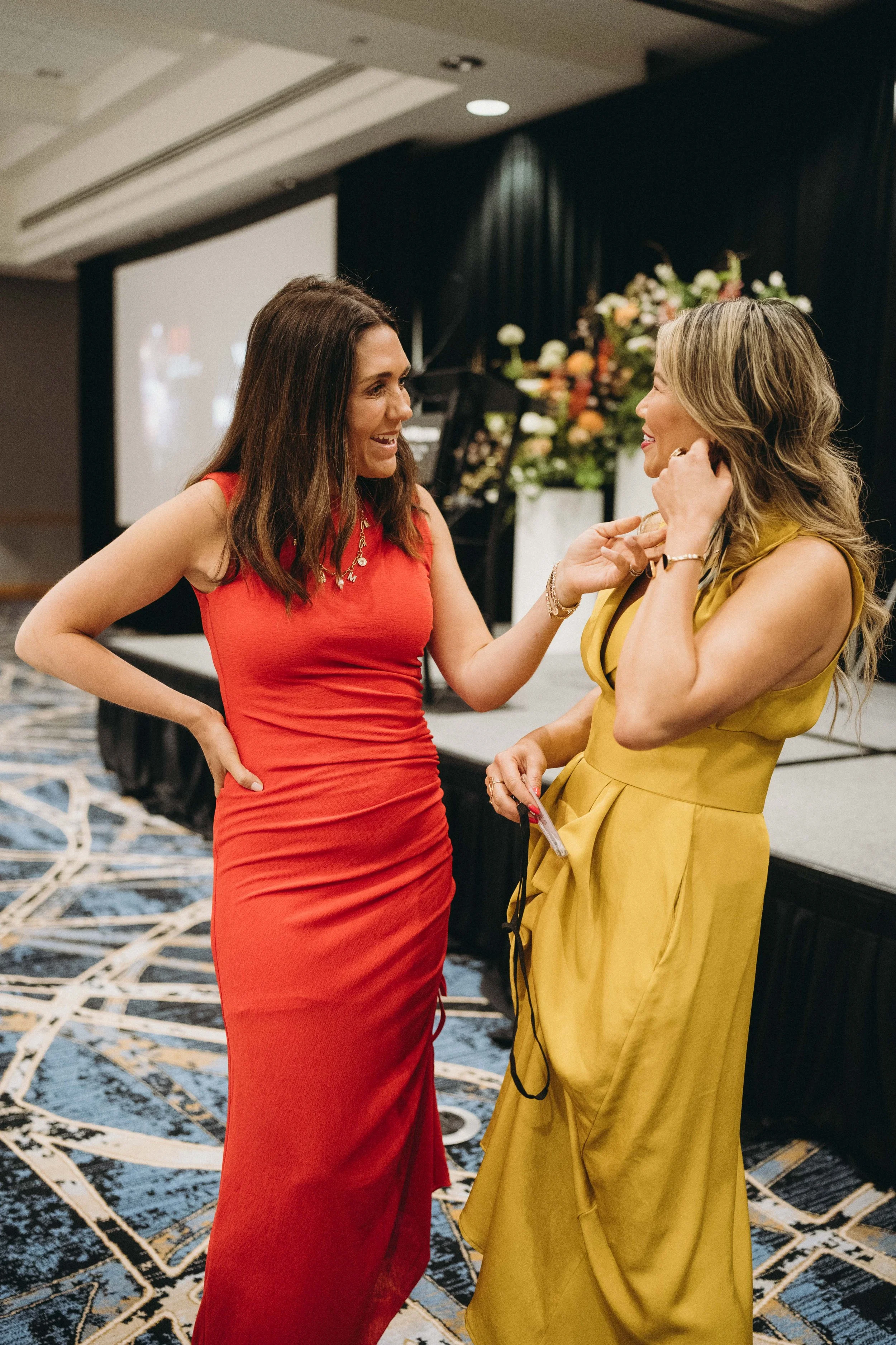 Two women standing and smiling at each other in a conference room. One woman is wearing a red dress with a gold necklace, and the other woman is wearing a yellow dress. There are flowers and a large white screen in the background.