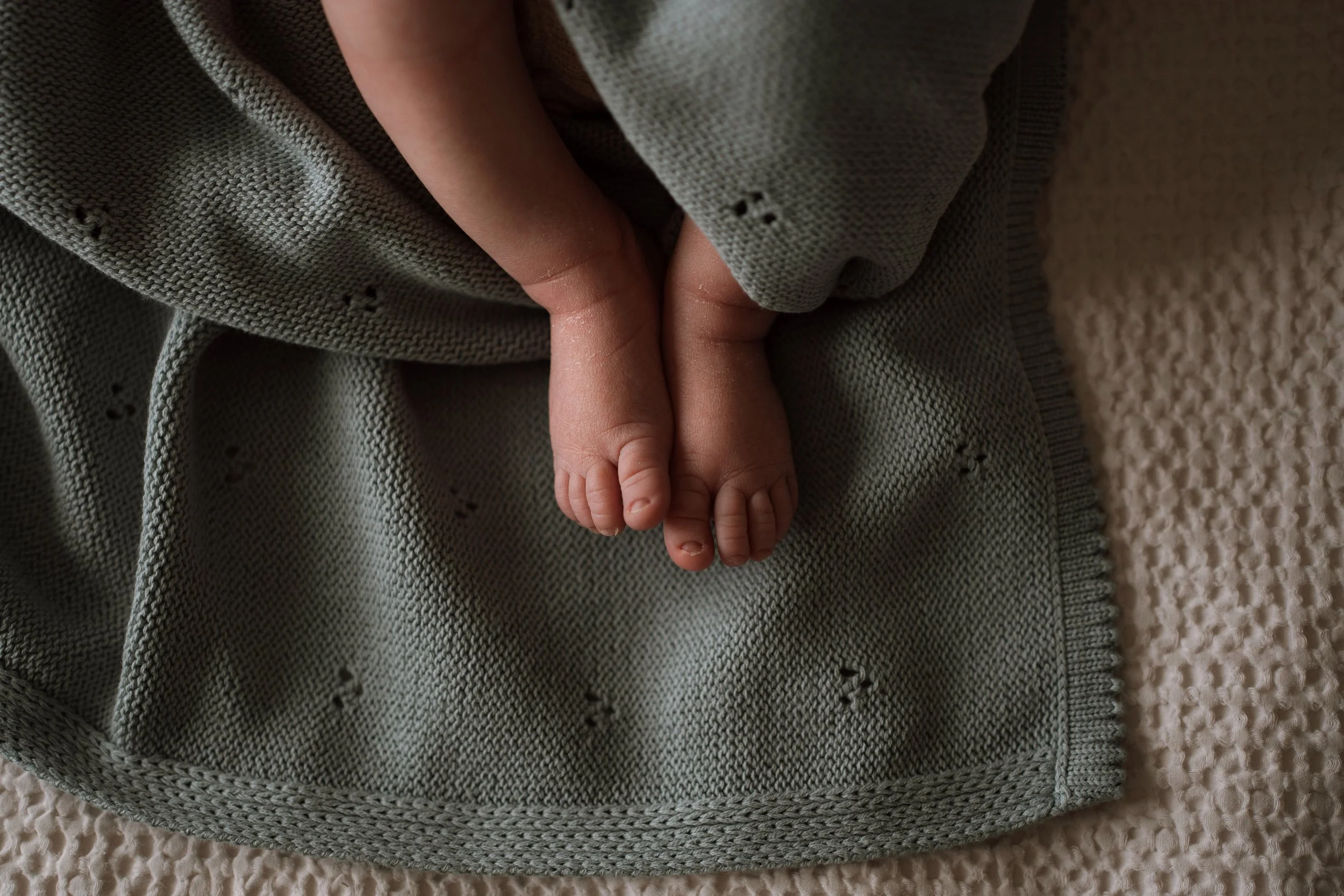 Close-up of a baby's tiny foot peeking out from under a gray blanket with embroidered holes, resting on a textured beige surface.
