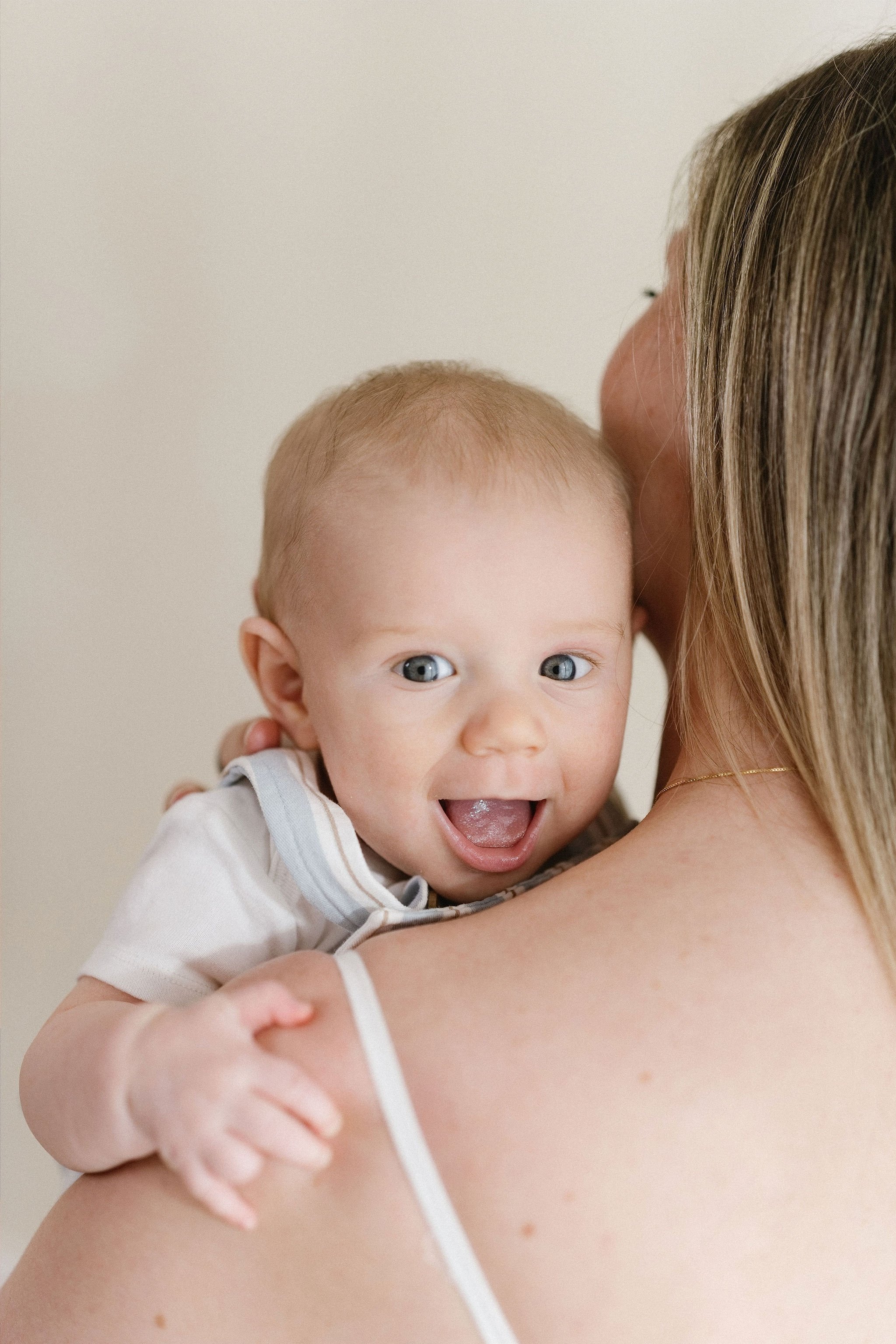 A woman holding a smiling baby with blue eyes and light hair, close to her shoulder, against a plain beige background.
