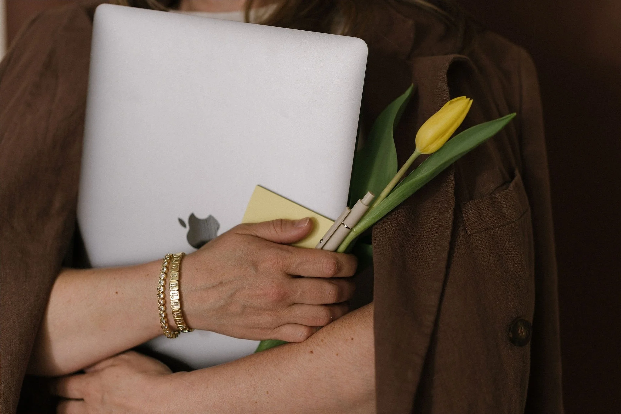 Person holding a MacBook, a yellow tulip, and a pad of sticky notes, wearing jewelry including pearl and gold bracelets.