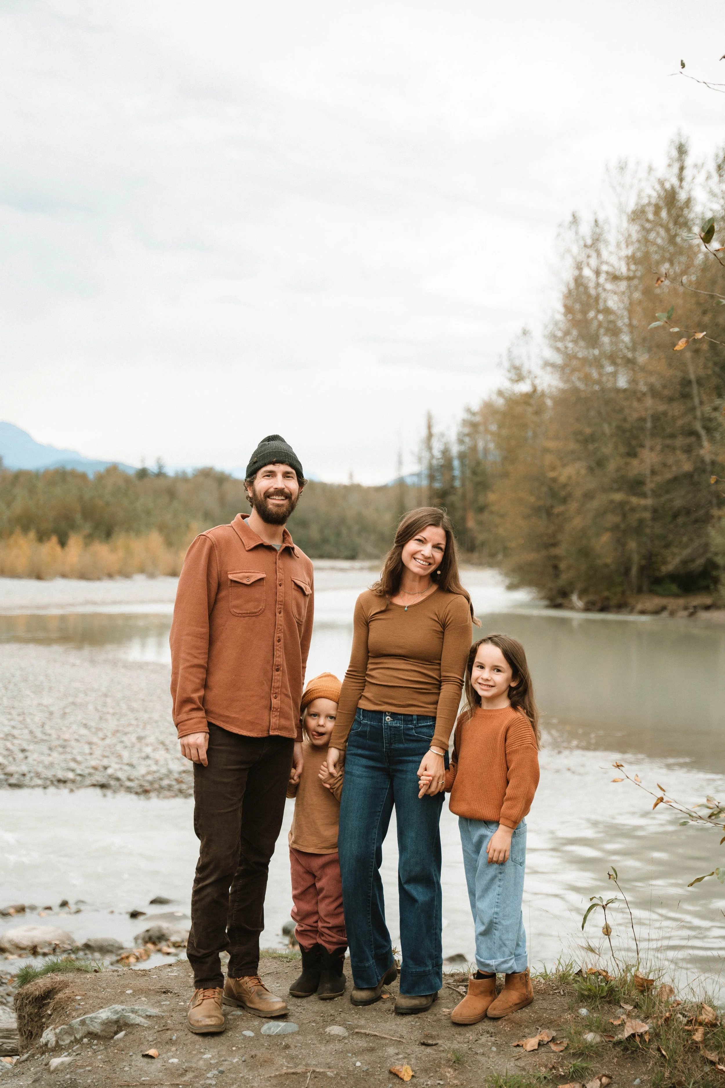 Family of four outdoors by a river, autumn setting, smiling, holding hands, casual clothing