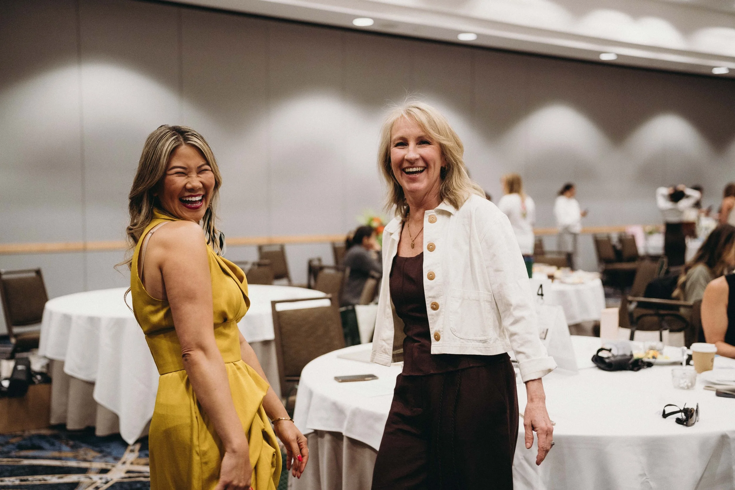 Two women smiling and standing in a conference room or banquet hall, with tables and chairs in the background, some people seated and some engaging in conversation.