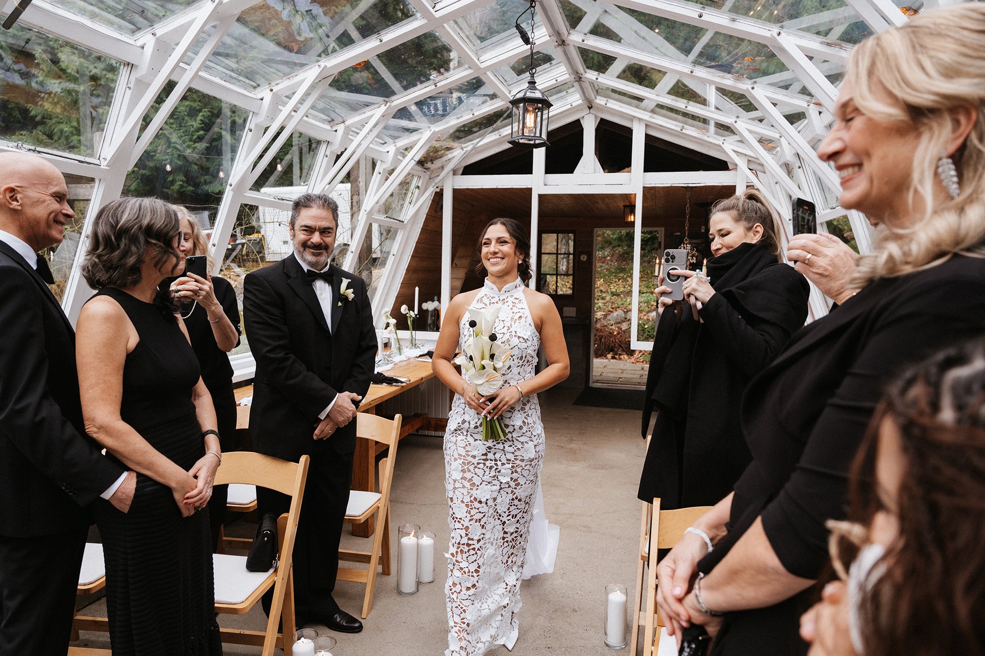 A bride in a white lace wedding gown is standing indoors, surrounded by smiling guests dressed in formal attire, during a wedding celebration in a glass-walled room with wooden accents.