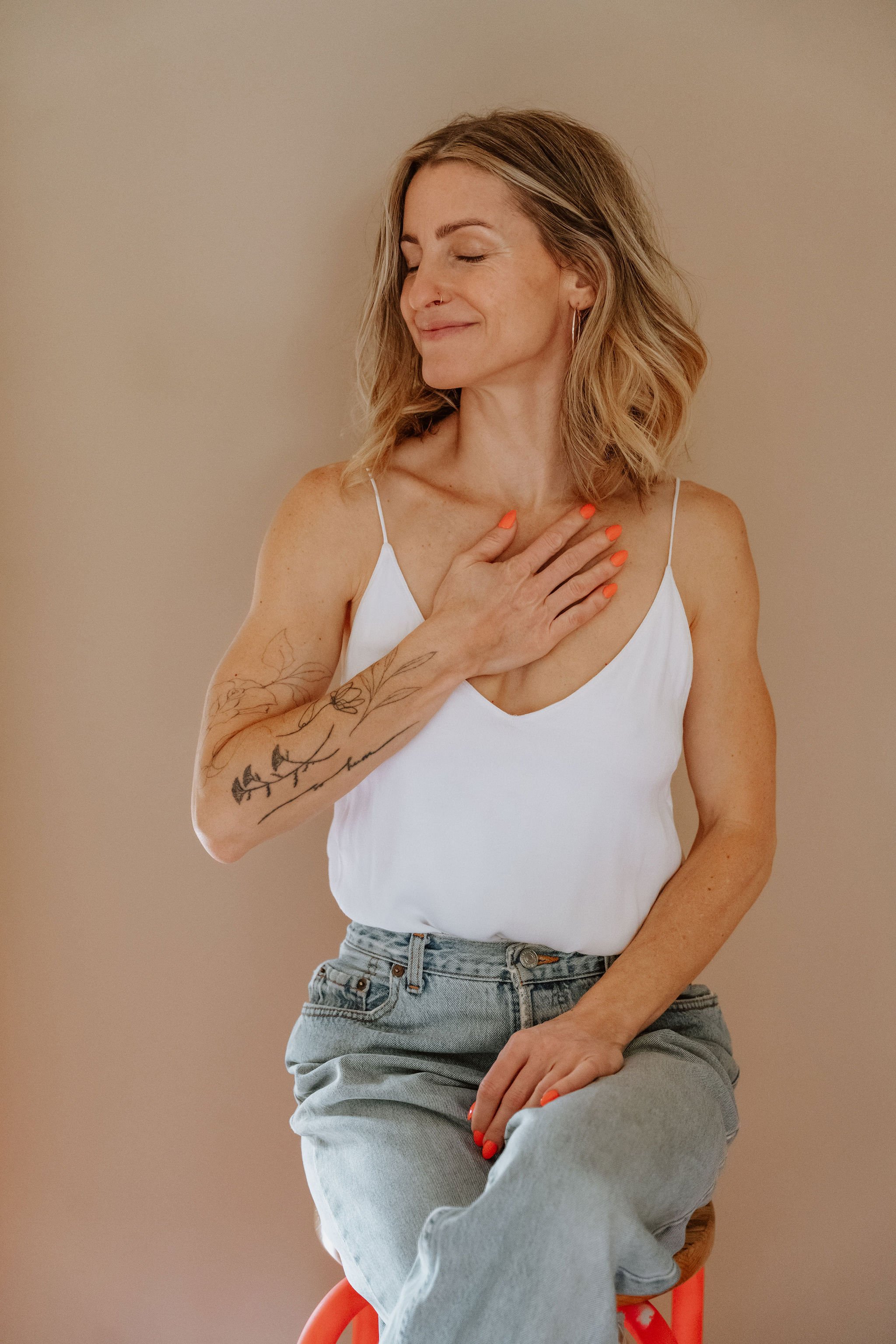 A woman with shoulder-length wavy hair, wearing a white spaghetti strap top and light blue jeans, sitting on an orange chair, with one hand on her chest and the other resting on her knee, smiling with eyes closed against a plain beige background.