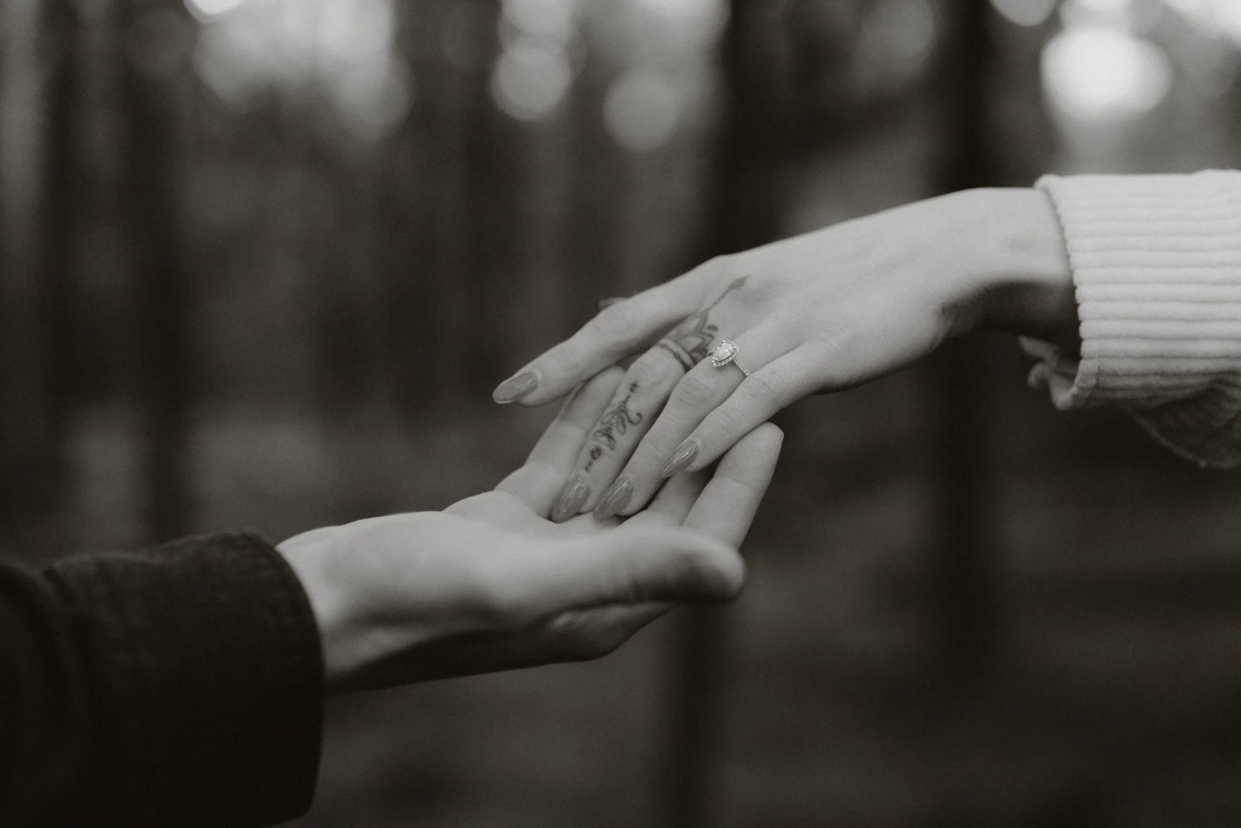 Close-up of two hands holding each other, one with a ring on the finger and a tattoo on the hand, in a black and white photo.