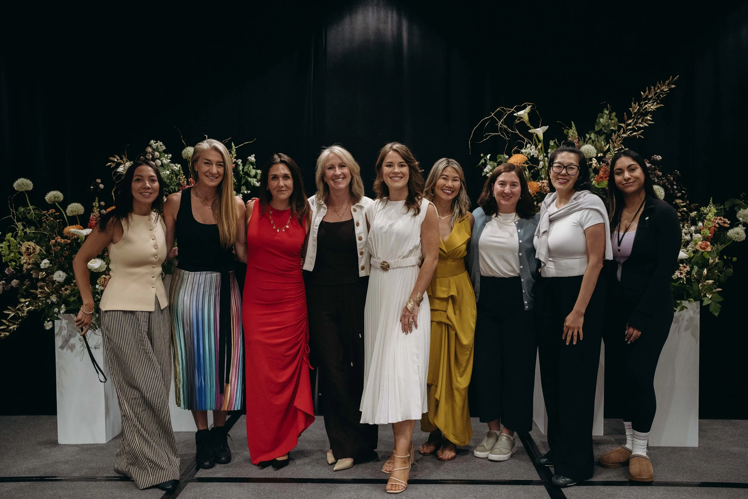 A group of nine women standing together in front of a black backdrop and floral arrangements, smiling and posing for a photo.