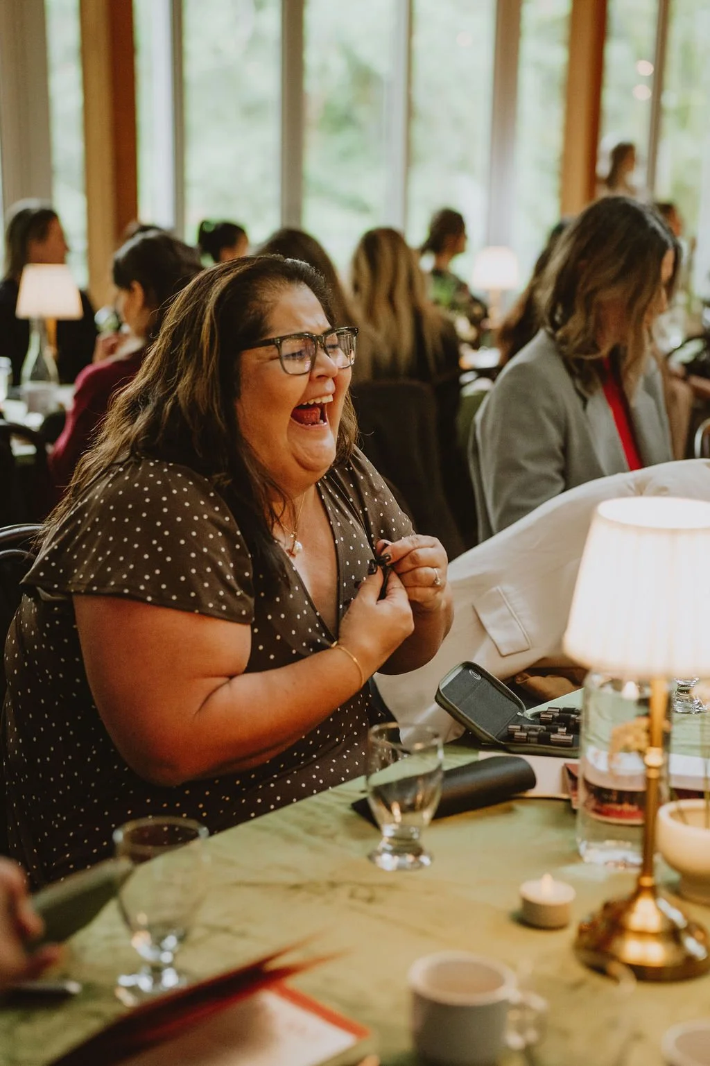 A woman with glasses, long dark hair, and a polka-dot dress is laughing with her hands clenched near her chest. She is seated at a table with glasses, a candle, and a lamp, with other people in the background in a warmly lit room with large windows.