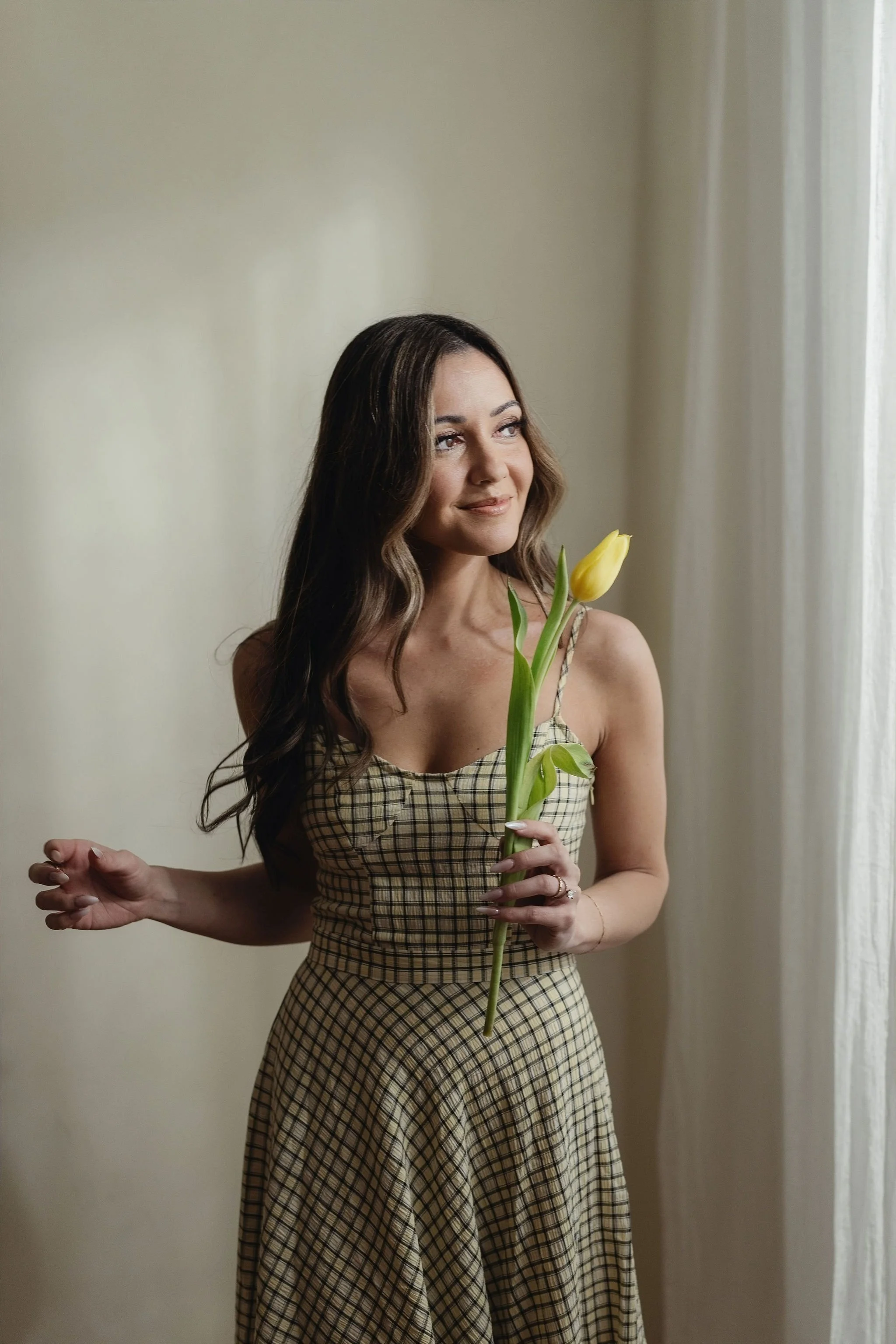 A woman with wavy brown hair holding a yellow tulip flower in front of her, standing in front of a plain beige wall with a light-colored curtain to her right, wearing a checkered dress and looking slightly to her left with a gentle smile.