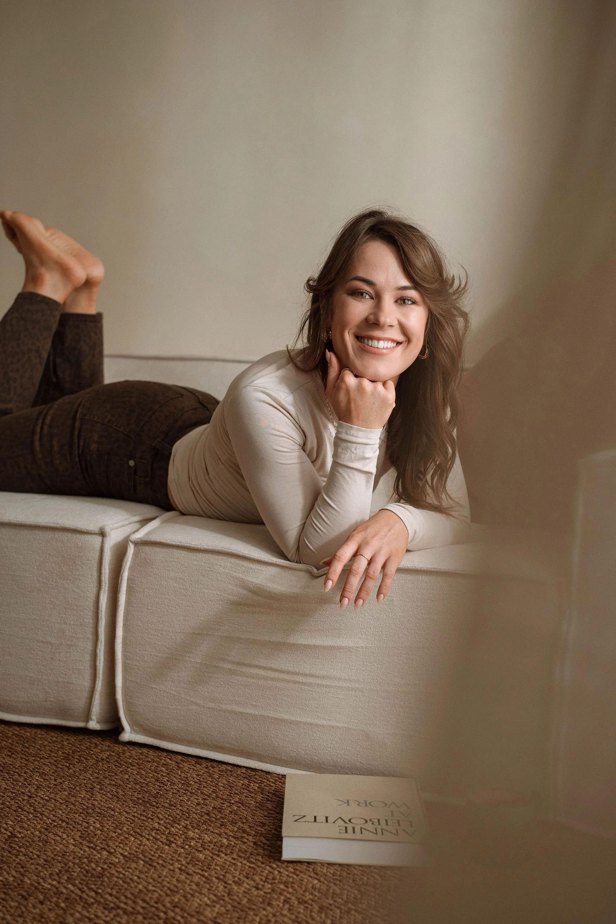 A woman with long brown hair and a white shirt is lying on her stomach on a beige couch, smiling at the camera, with her head resting on her hand and her knees bent up behind her. A book titled "Aline Levezzz, Le Livre de Work" is on the floor near t