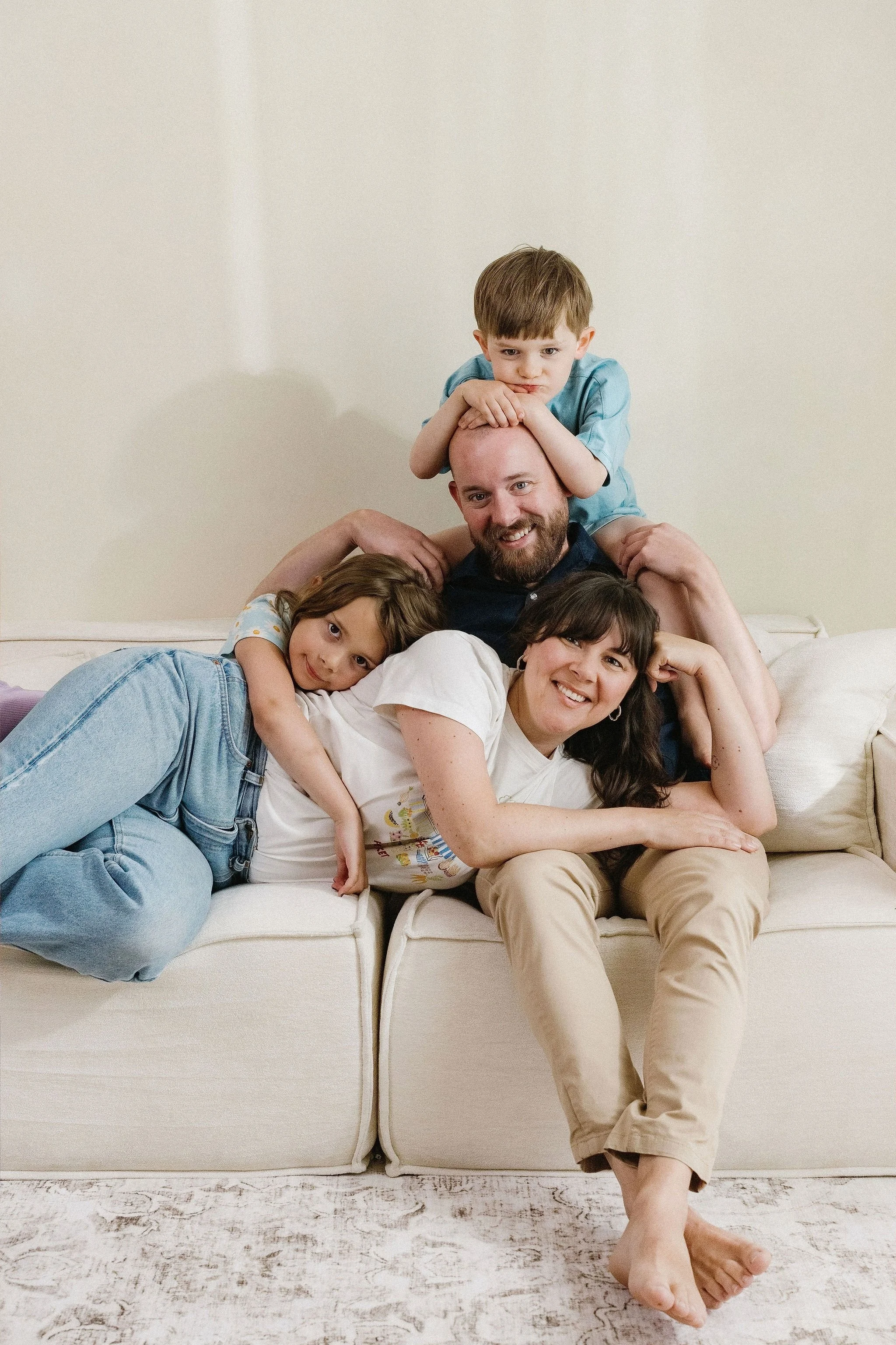 A happy family of four lying on a cream-colored sofa in a living room. The mother and daughter are on the left, with the daughter laying on her side and the mother smiling and resting her head on her hand. The father is lying behind them, smiling, wi