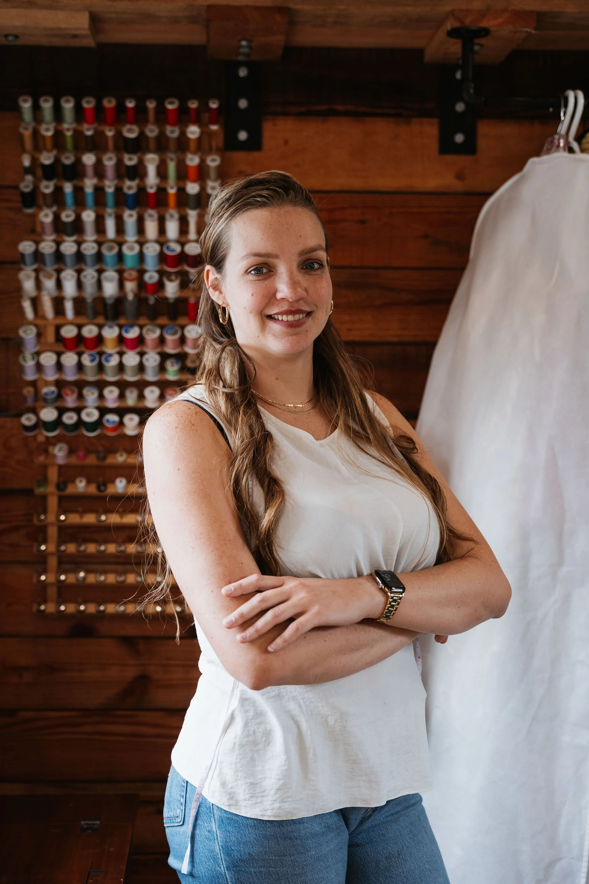 A young woman with long wavy brown hair and fair skin stands with her arms crossed, smiling in front of a wall display of colorful threads for sewing or embroidery, inside a wooden-paneled room.