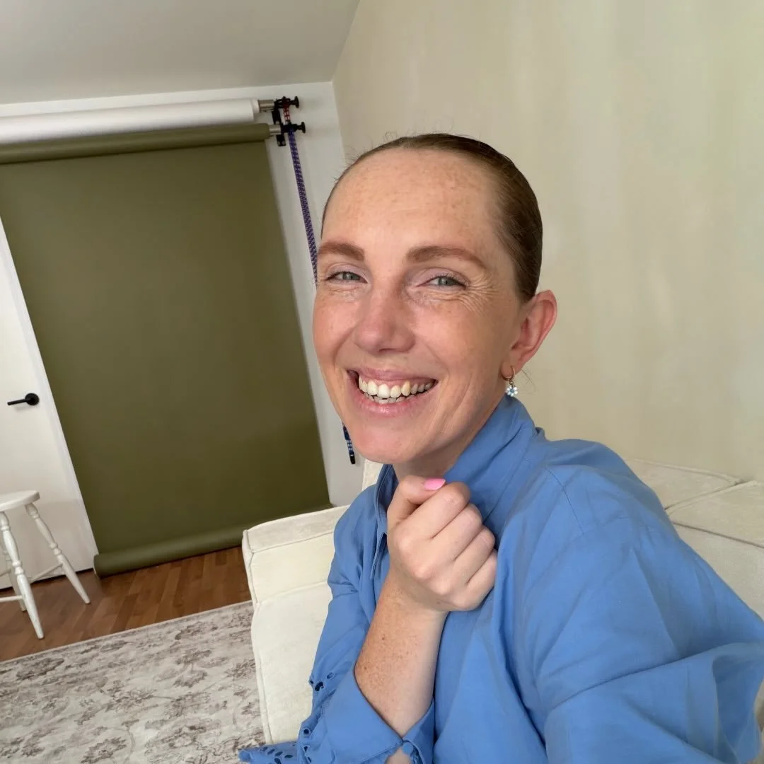 Smiling woman with light skin, red hair tied back, blue shirt, sitting on a white couch in a living room with a green backdrop curtain, a small white chair, and a wood floor.