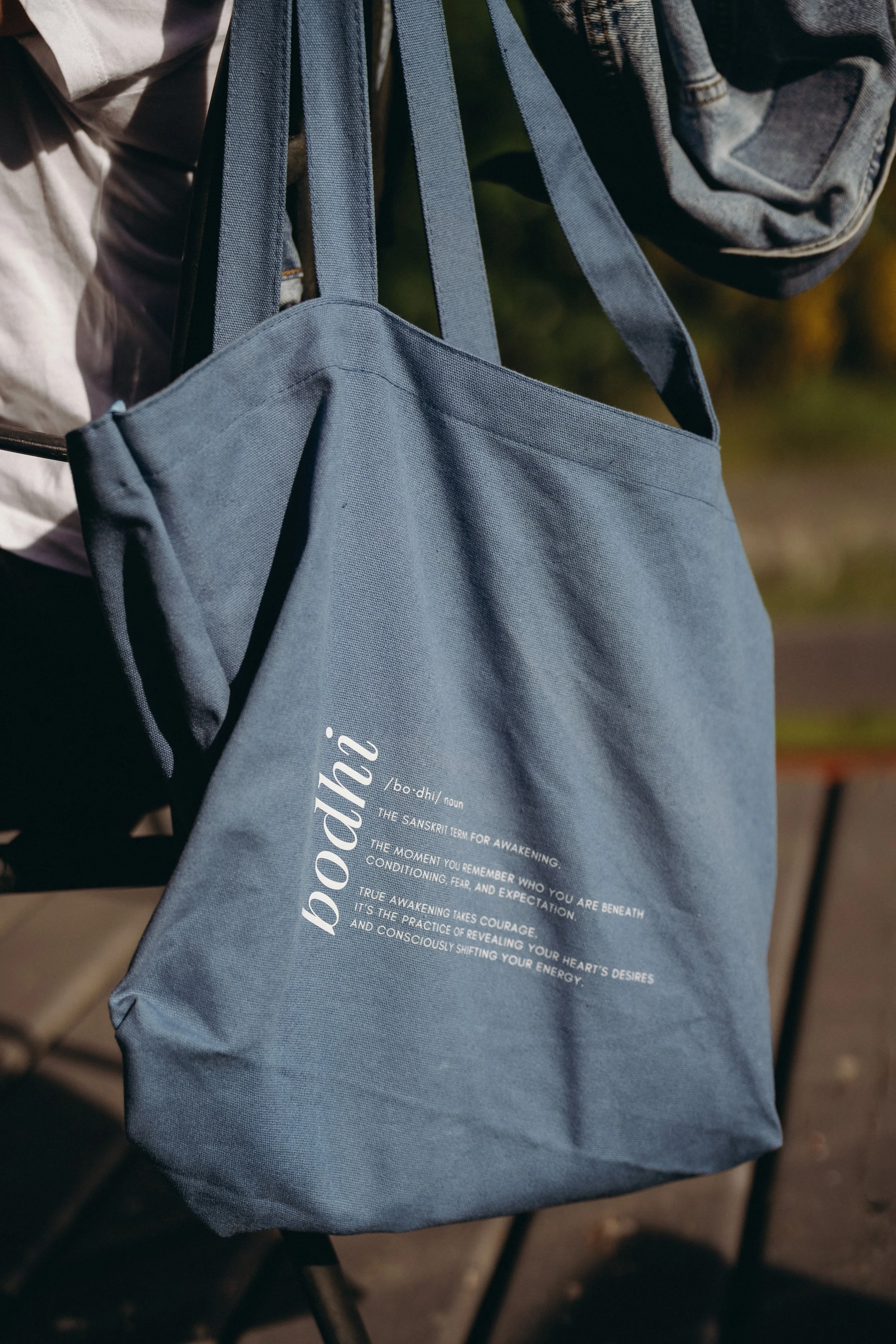 A blue tote bag hanging from a person's shoulder, with white text and definitions printed on it. The background shows part of a person wearing a denim jacket and a white shirt.