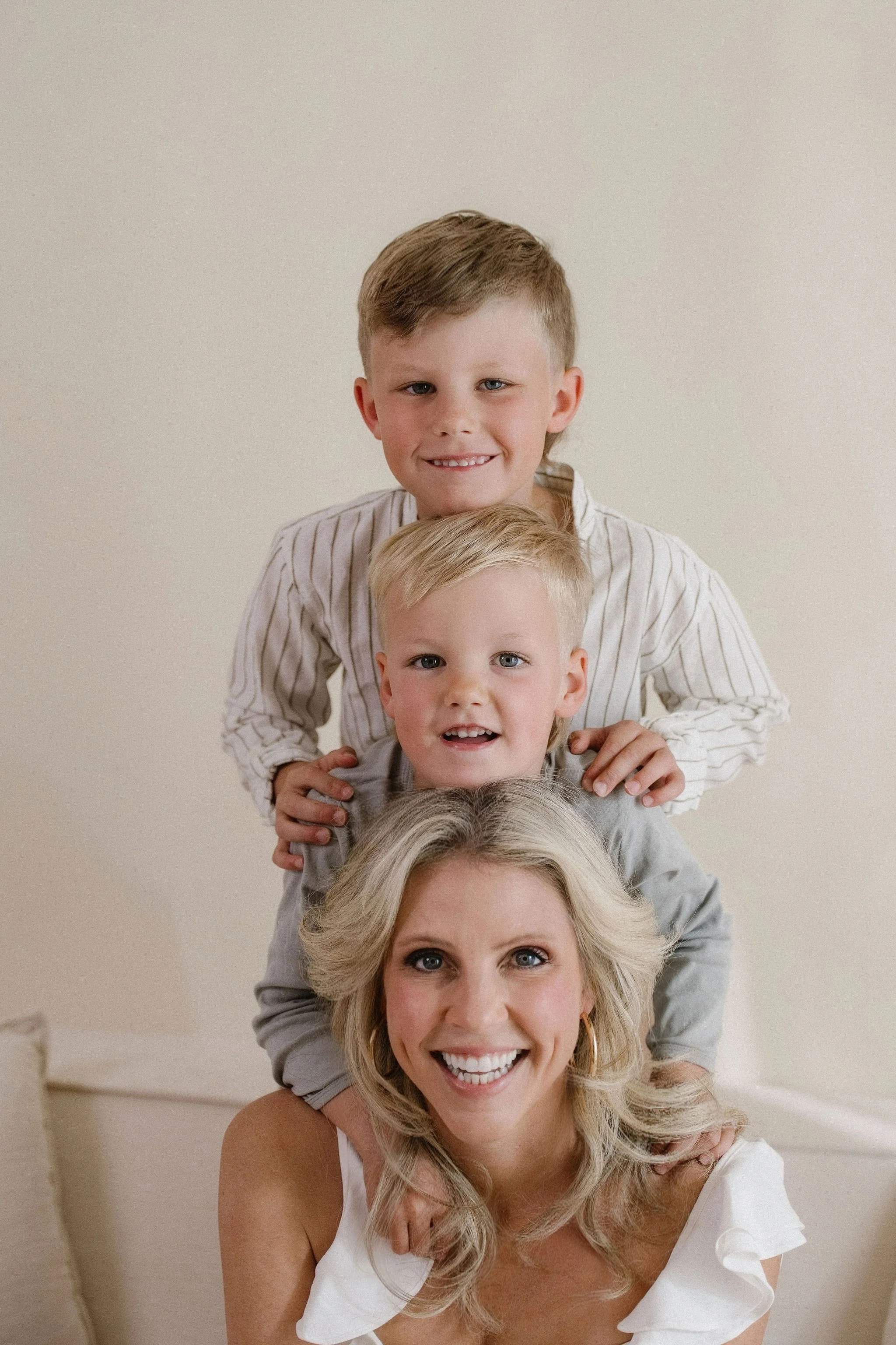 A woman with blonde hair smiling and four children stacked vertically, with each child on the shoulders of the one below, against a plain neutral background.