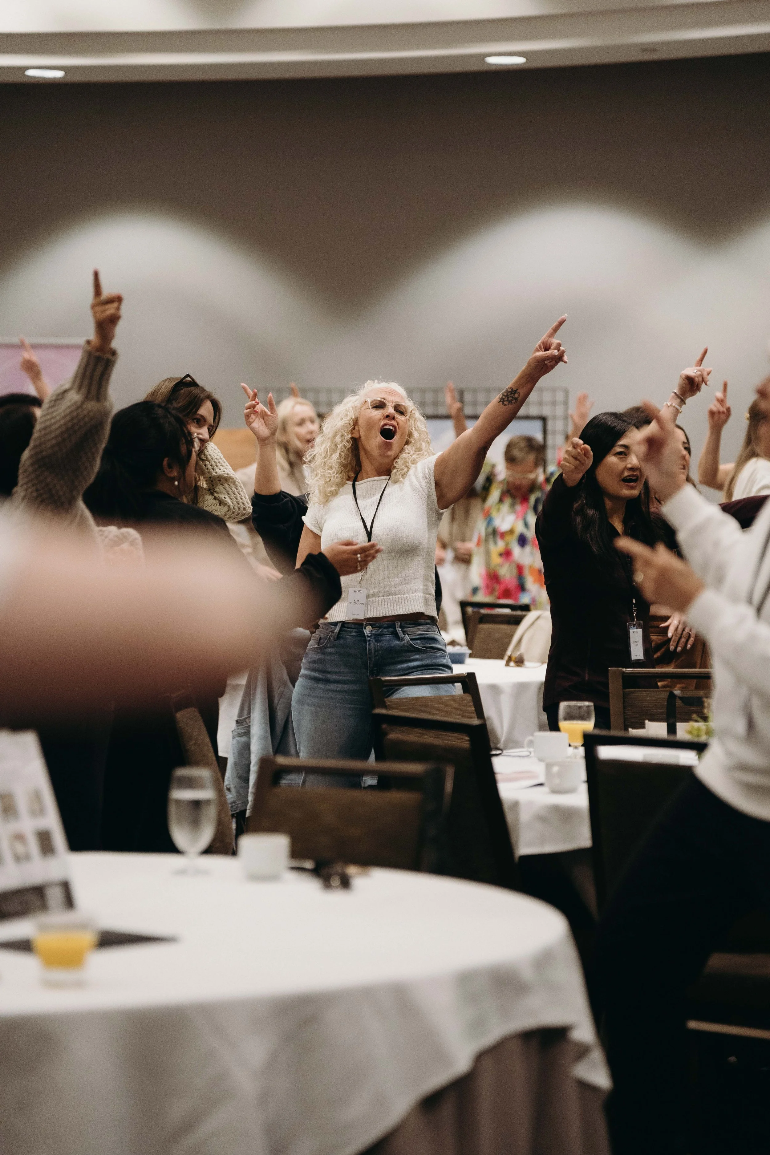 A group of women singing and raising their arms in enthusiasm at a conference or gathering, with tables, chairs, and breakfast items visible in the foreground.