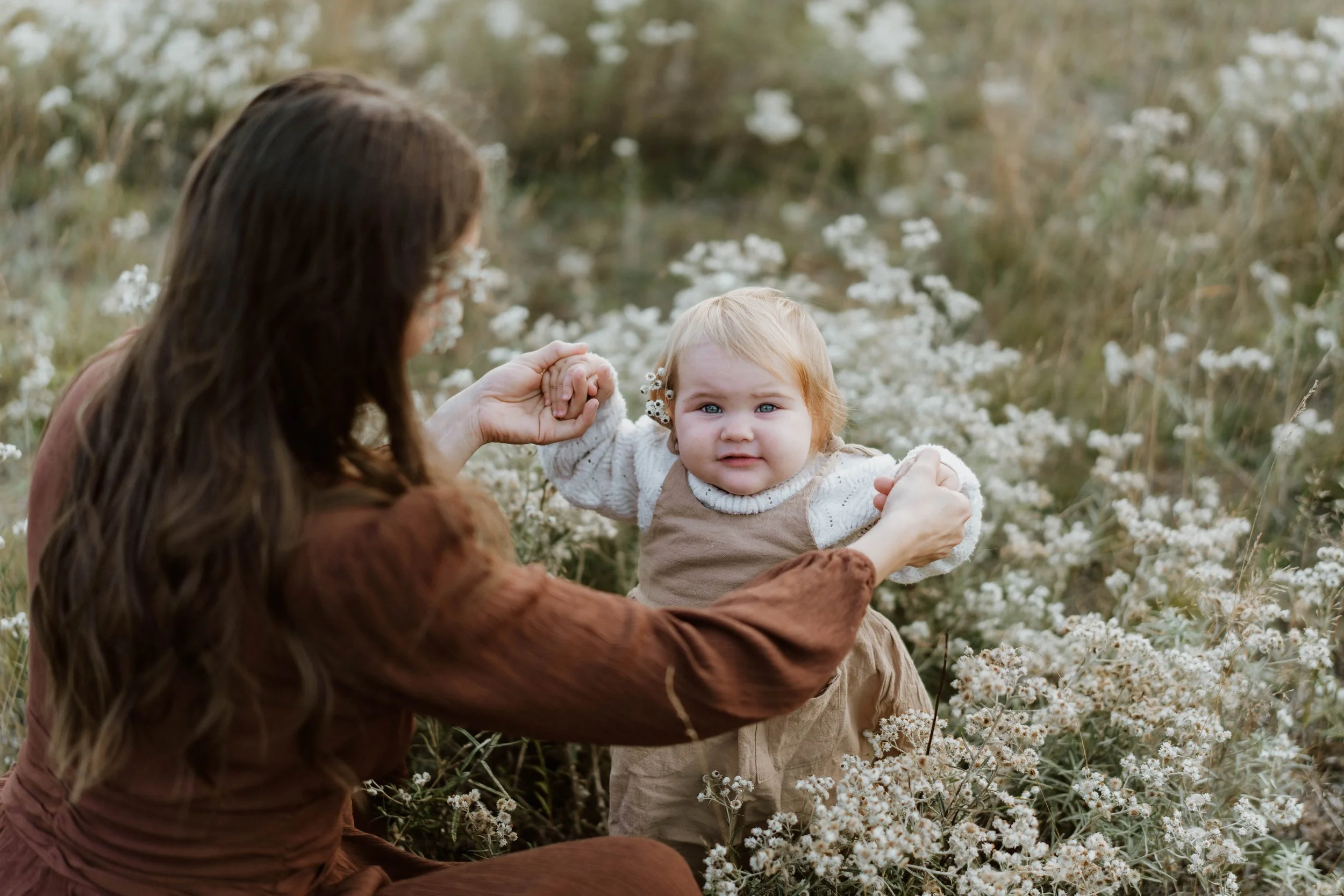 A woman with long dark hair helping a young girl stand in a field of white wildflowers, with the woman holding the girl's hands. The girl has blonde hair, blue eyes, and is wearing a beige dress with puffy sleeves, looking at the camera.