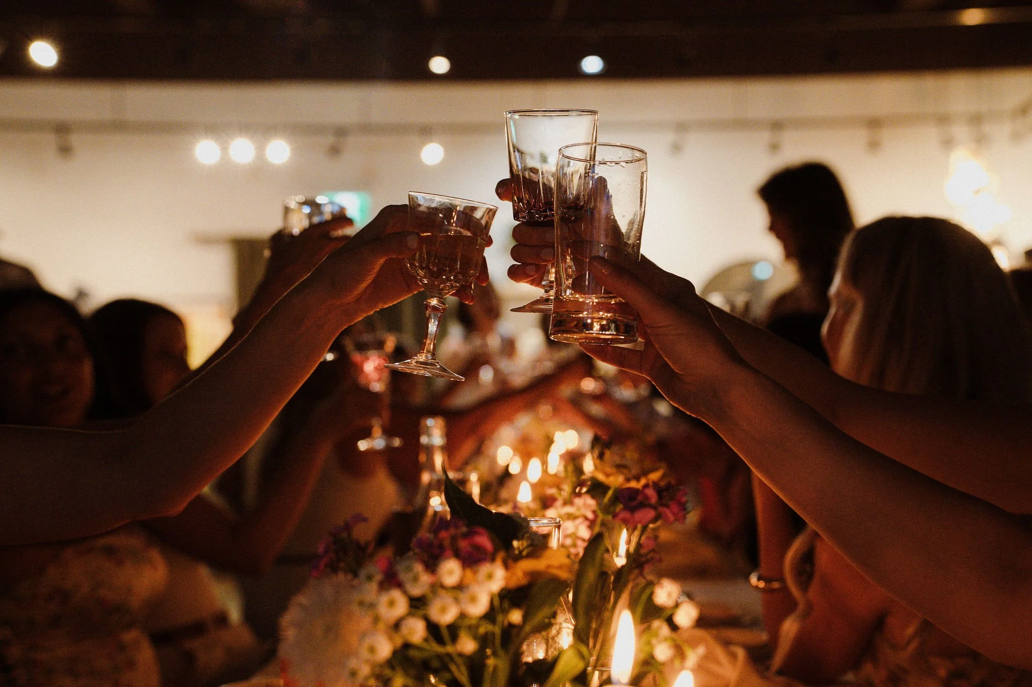 People raise glasses for a toast at a celebration or gathering in a dimly lit room with floral centerpieces.
