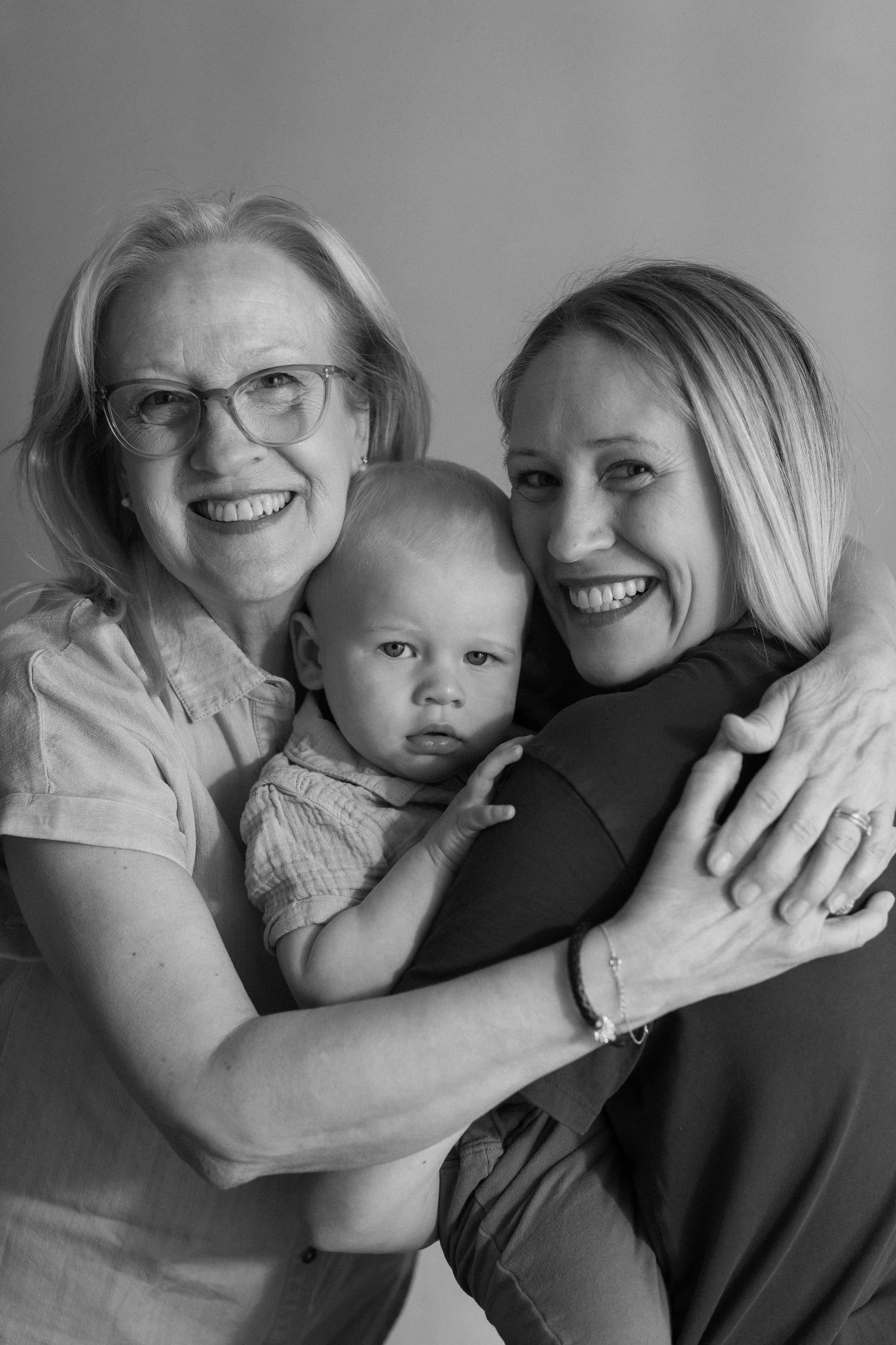 A black and white photo of three generations of women hugging each other tightly and smiling, with a young child in the center.