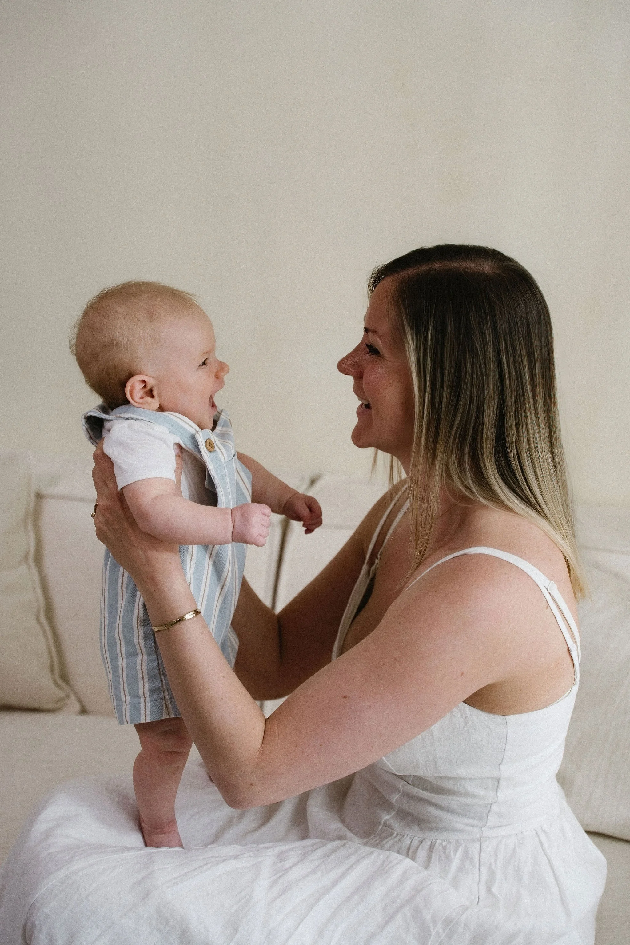 A woman holding a smiling baby boy in a bedroom with a white headboard and a beige wall behind them