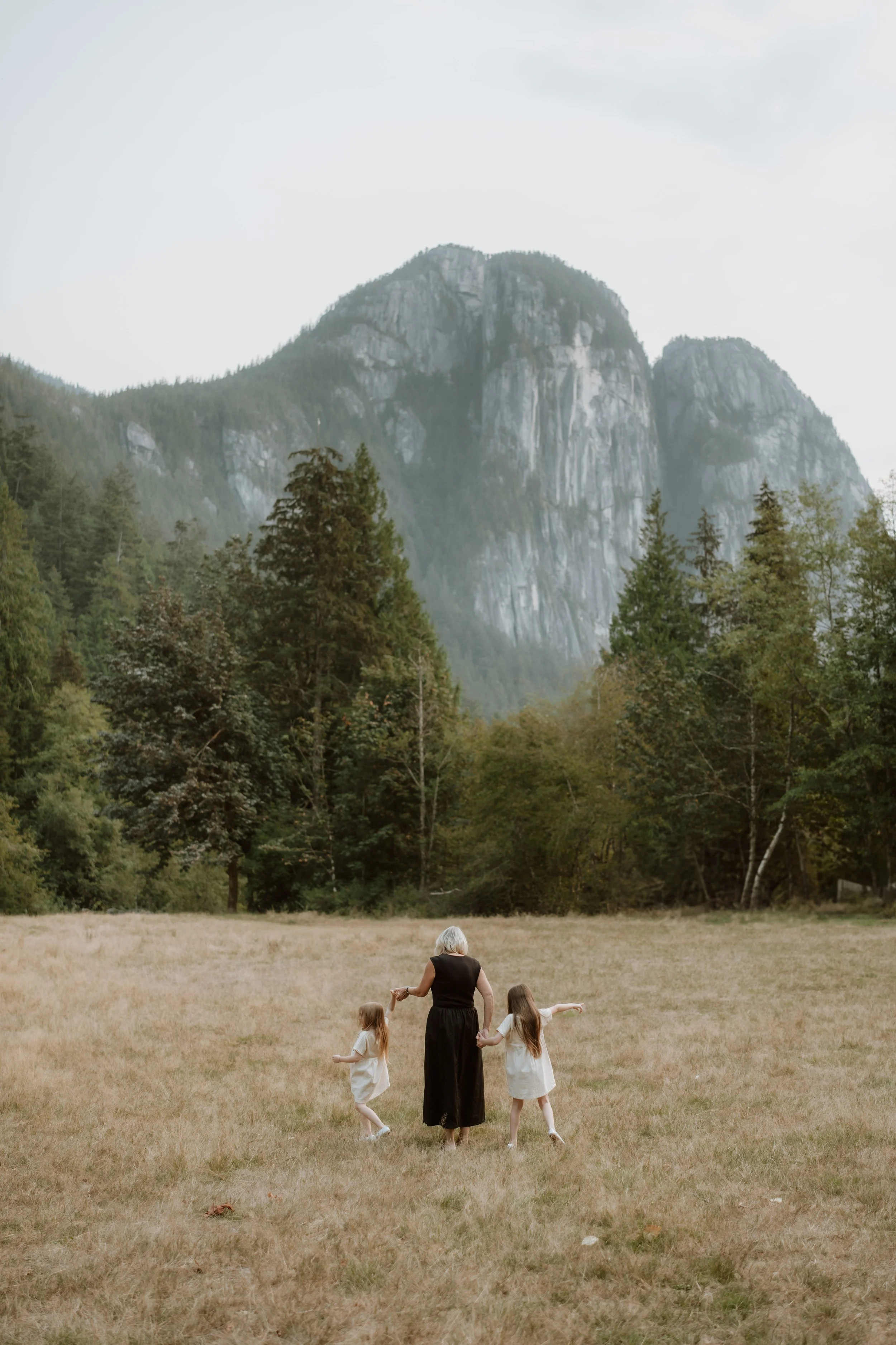 An older woman and two young girls walking hand-in-hand across a grassy field with mountains and trees in the background.