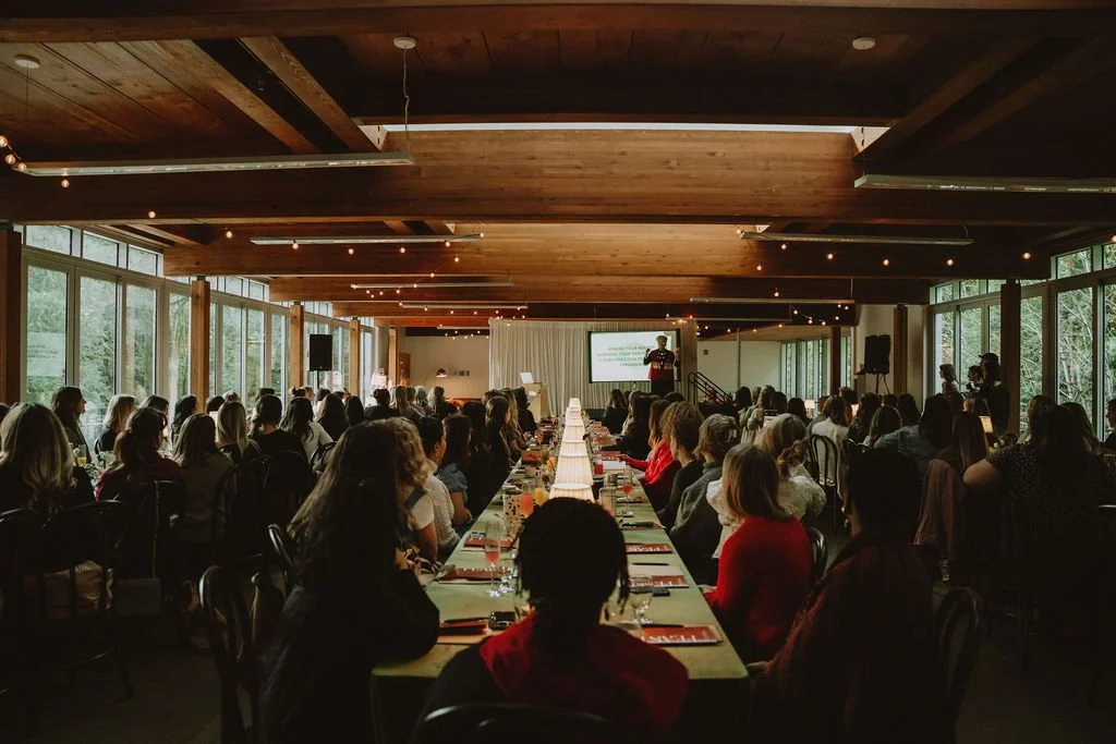 A large conference or seminar room filled with people seated at long tables, listening to a speaker on stage in front of a screen. The room has large windows and a wooden ceiling with string lights.