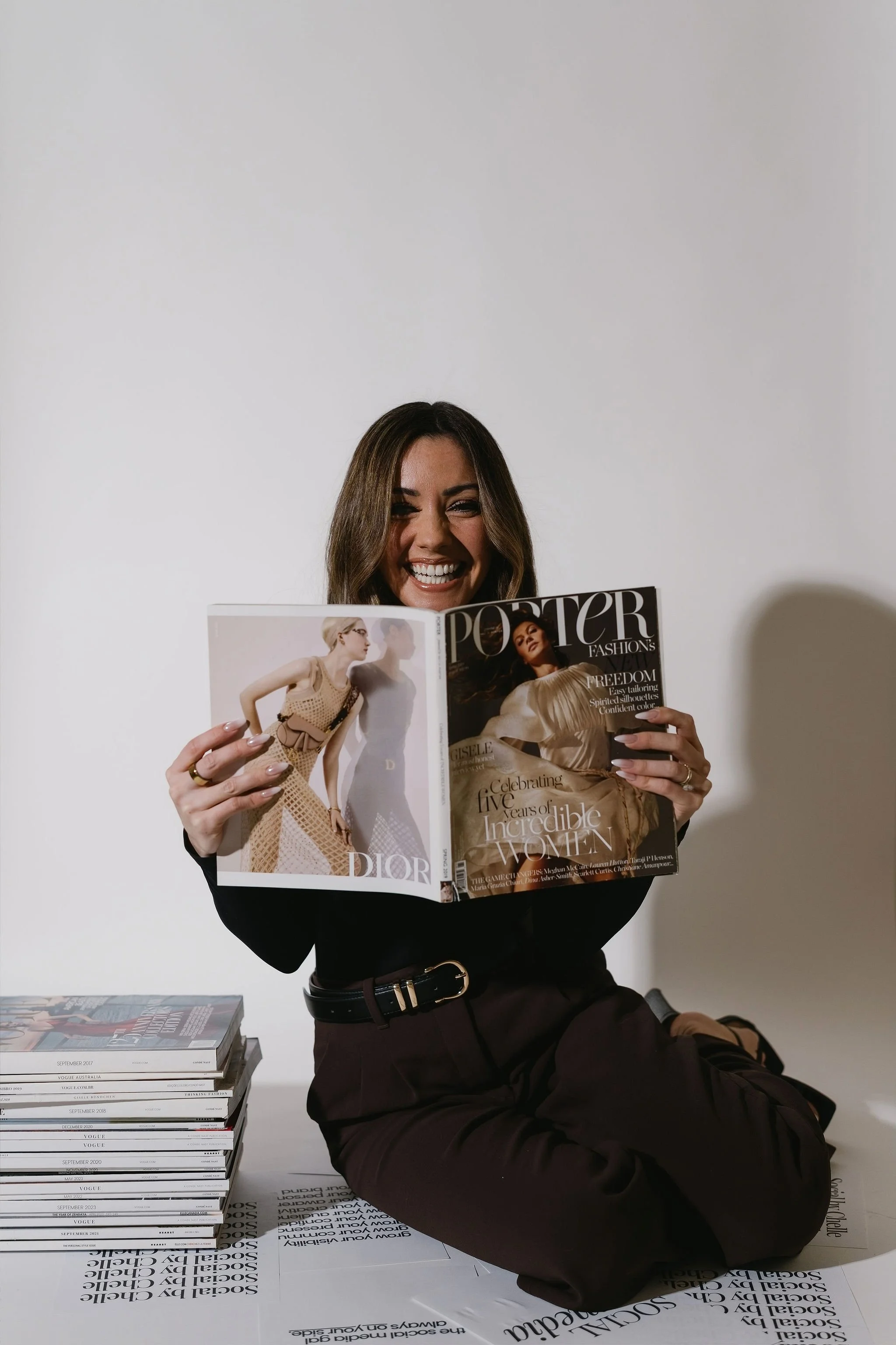 A woman with long brown hair smiling and sitting on the floor, holding an open fashion magazine with models on the pages. She is wearing a black top and dark pants. There is a stack of magazines on the table beside her and a white table covering with